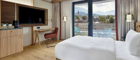 A hotel room with a large bed, a desk with a chair, a television, and a view of the city through a large window.
