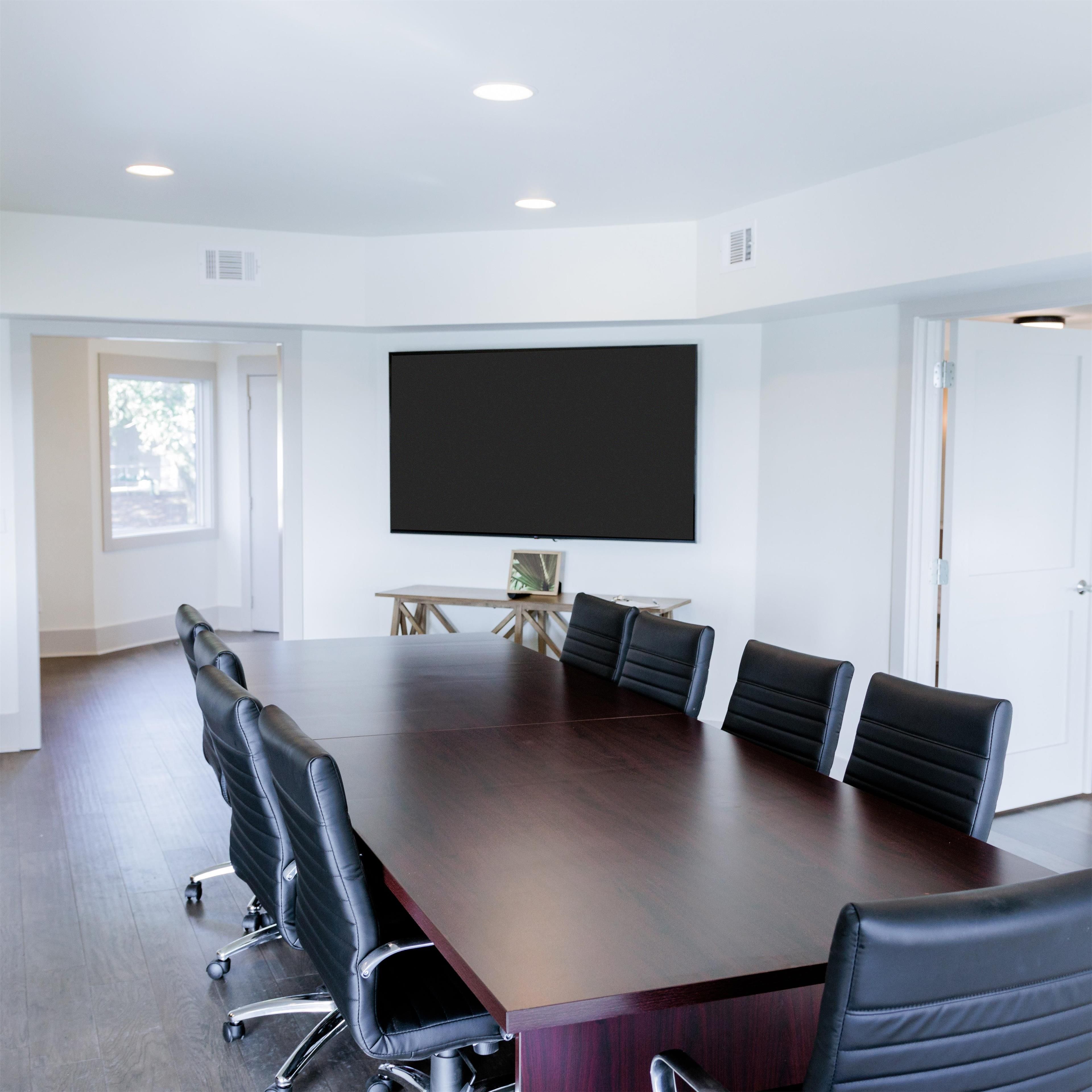 Une salle de conférence dotée d’une grande table en bois foncé, de six chaises en cuir noir, d’un grand téléviseur à écran plat fixé au mur et d’une petite table en bois avec une lampe.