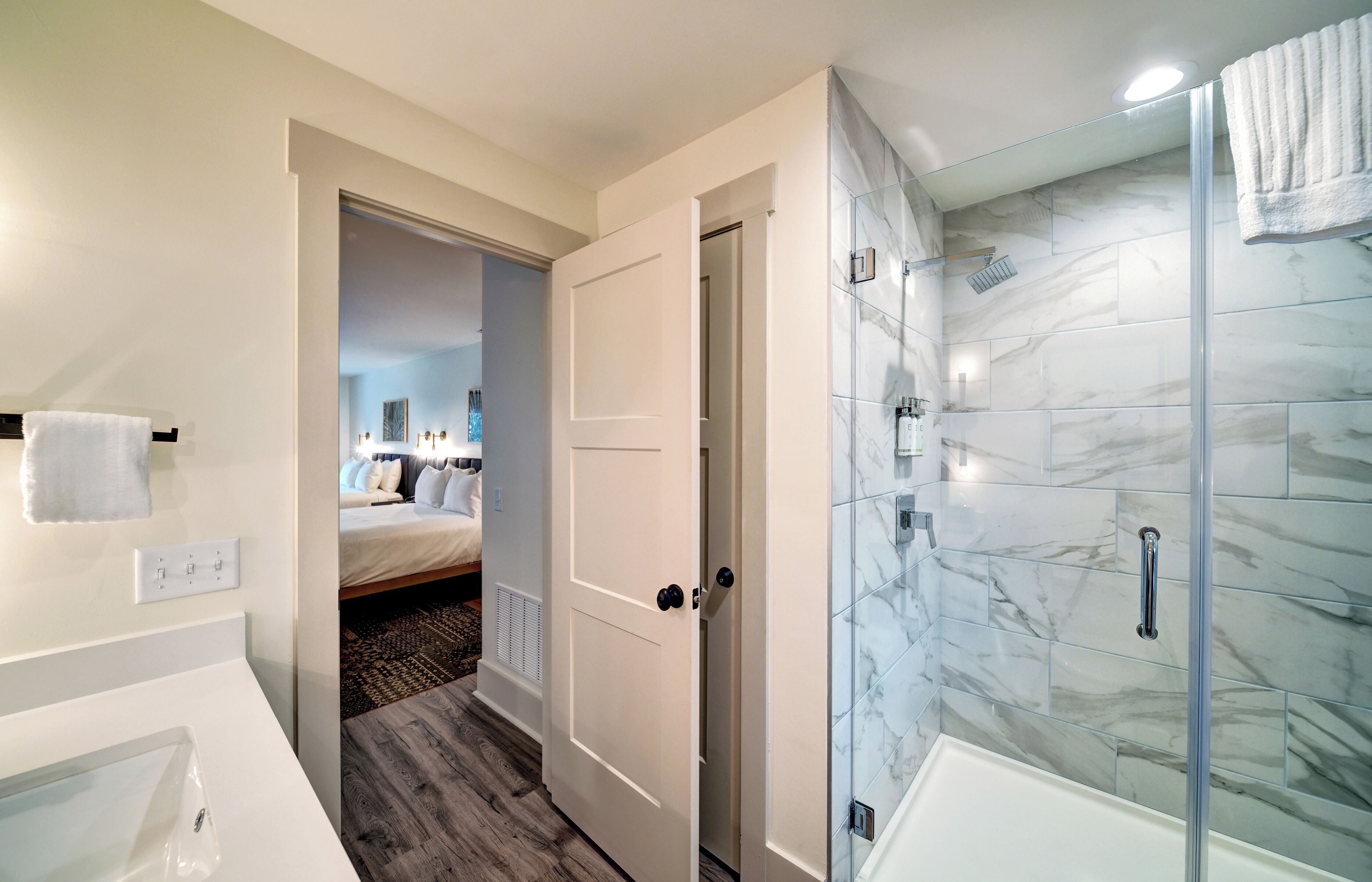 A modern bathroom with a glass-enclosed shower, a white bathtub, and a visible door leading to a bedroom.