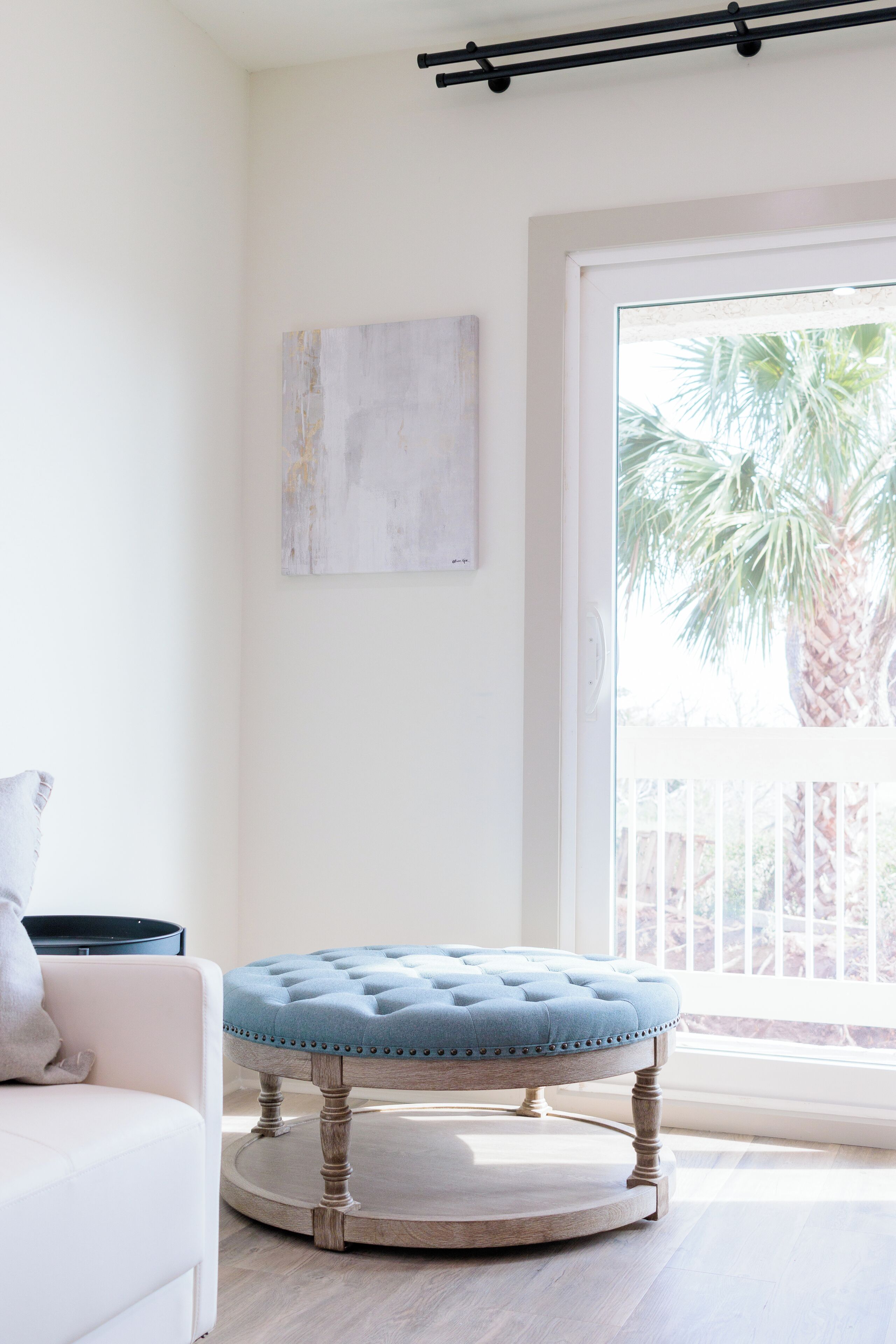 A room with a round wooden ottoman, a white sofa, and a large window with a view of palm trees.