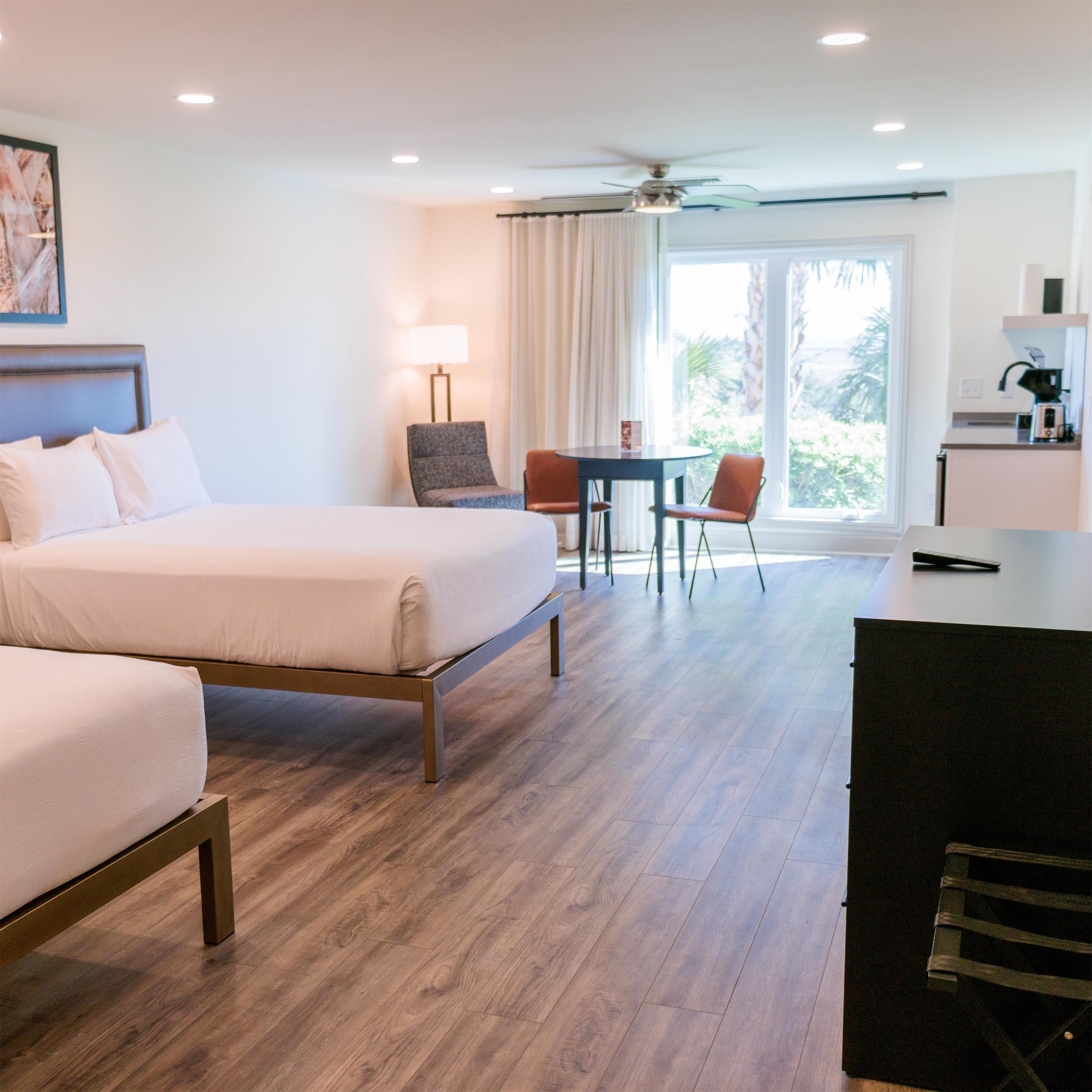 A hotel room with two beds, a desk, a chair, a table, and a view of the outdoors.