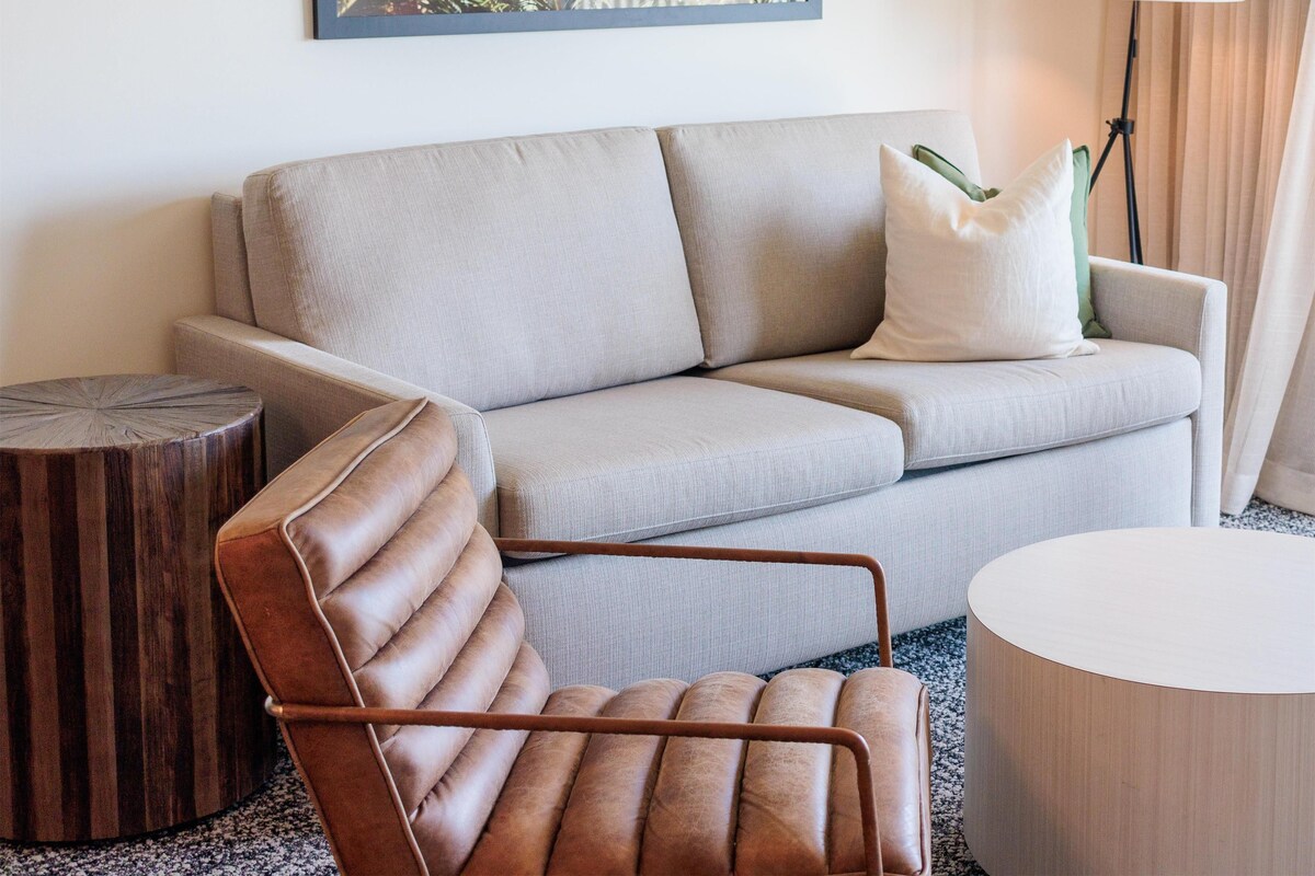 Una camera d'albergo moderna con un divano grigio, una poltrona in pelle marrone, un tavolino rotondo in legno, una lampada bianca e un quadro con motivi vegetali appeso al muro.