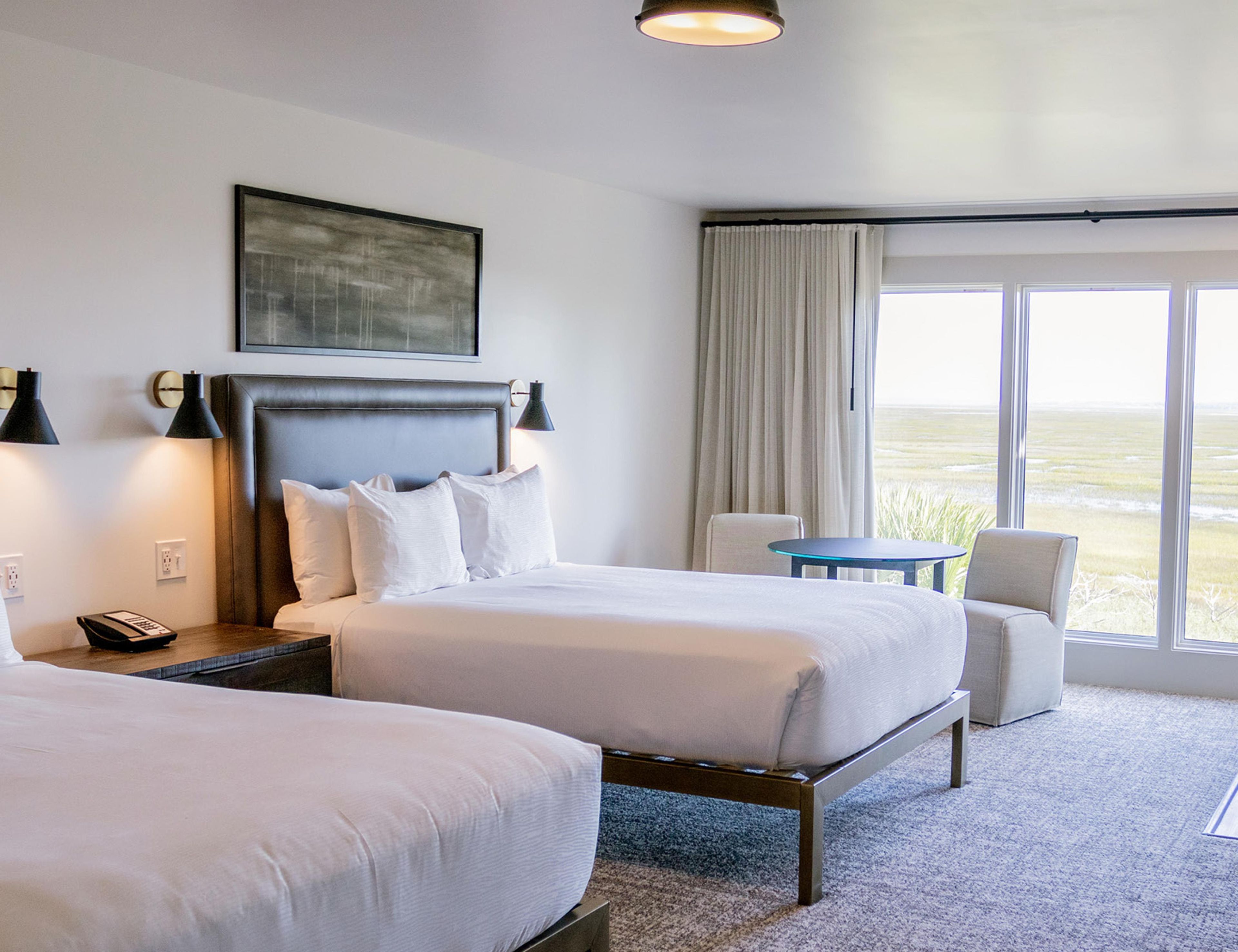 A hotel room with two beds, a desk, a chair, a window with a view, and a painting on the wall.