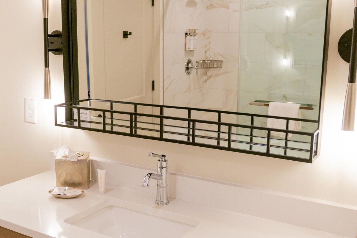 Un bagno con un lavandino bianco, uno specchio con cornice nera e uno scaffale in vetro sopra il lavandino.