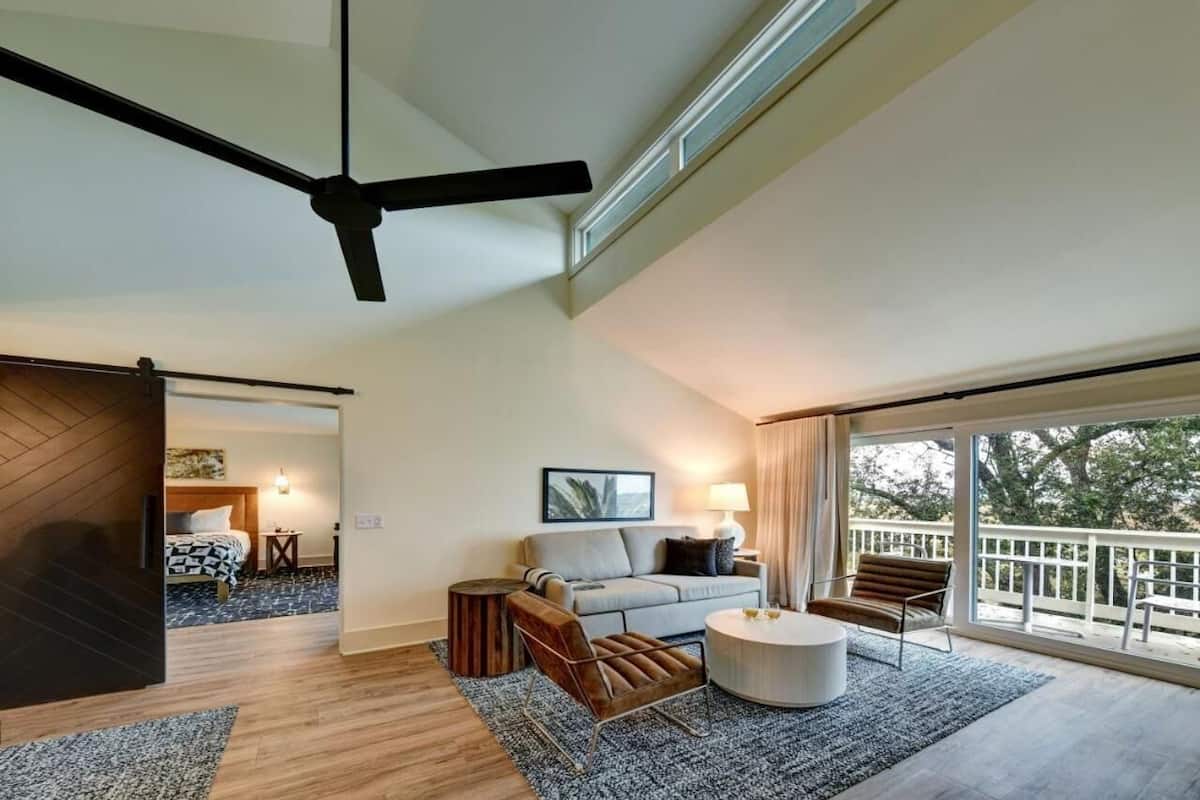 Una sala moderna con sofá, mesa de centro y ventilador de techo. Tiene un balcón con barandilla y vistas a árboles.