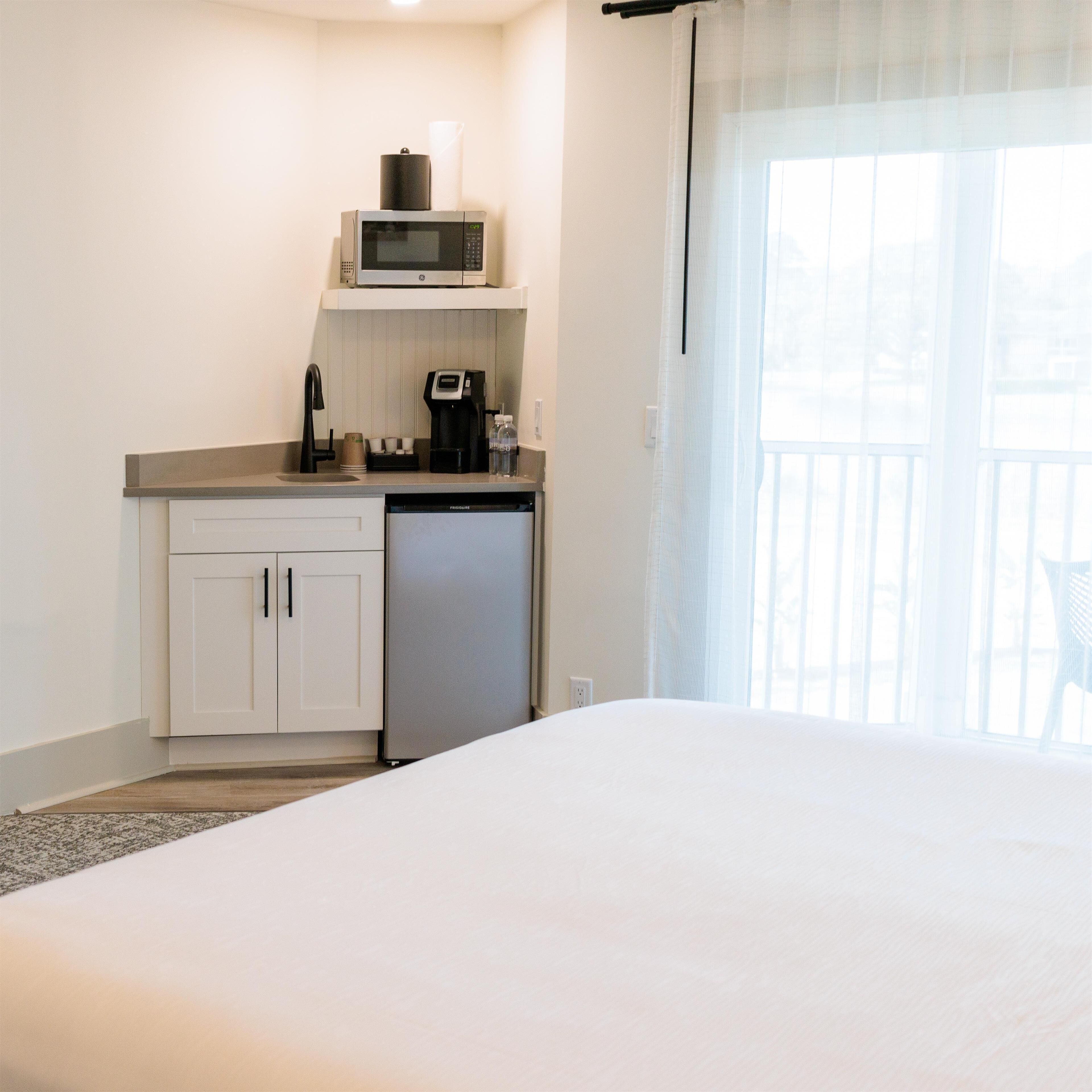 A hotel room with a bed, a kitchenette with a microwave, coffee maker, and dishwasher, and a balcony with a chair.