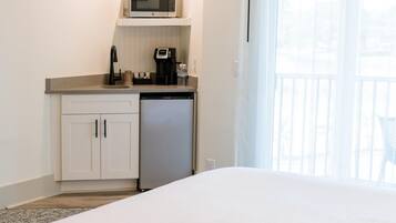 A hotel room with a bed, a kitchenette with a microwave, coffee maker, and dishwasher, and a balcony with a chair.