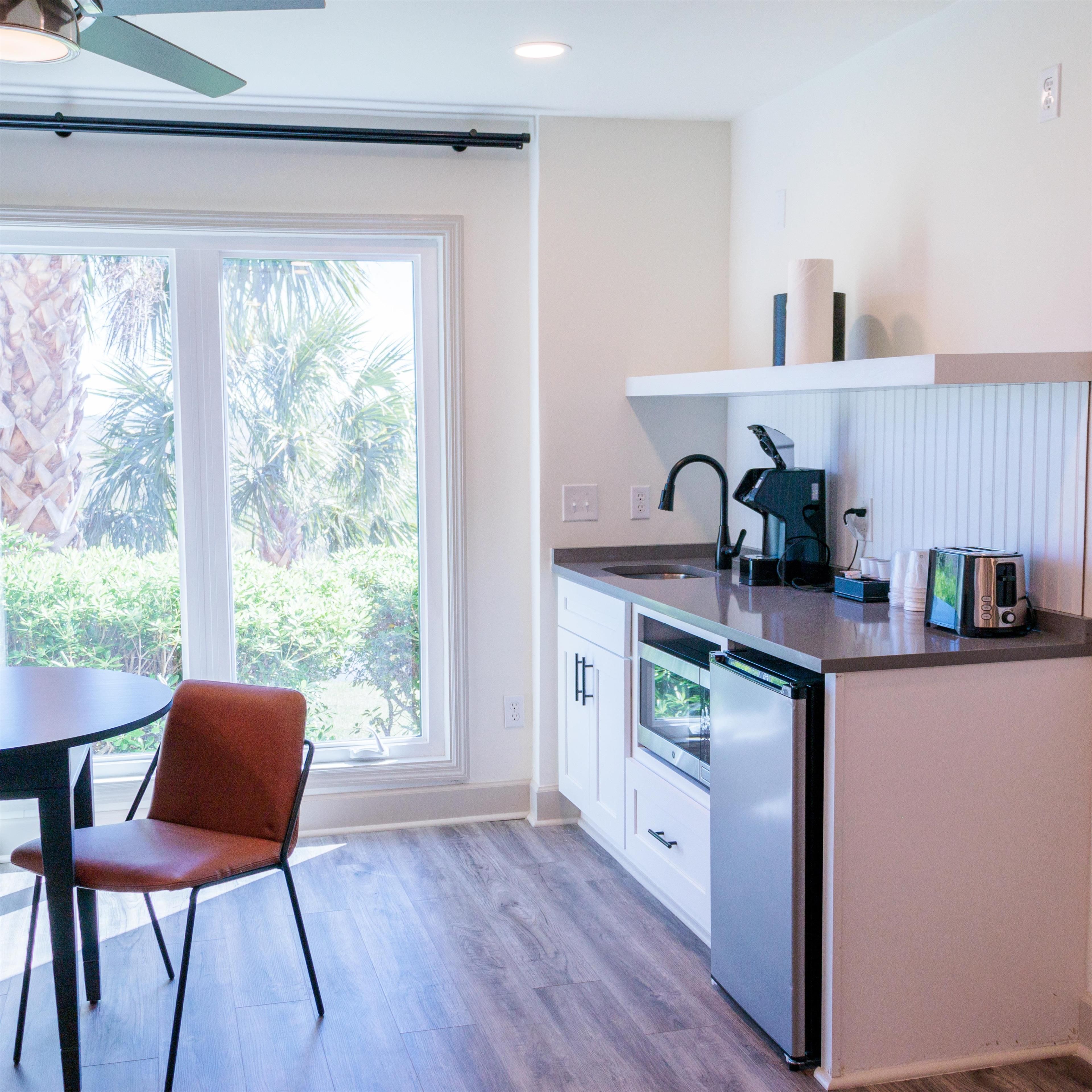 A modern kitchen with a built-in dishwasher, a coffee maker, and a toaster. There is a window with a view of palm trees and a chair in the dining area.