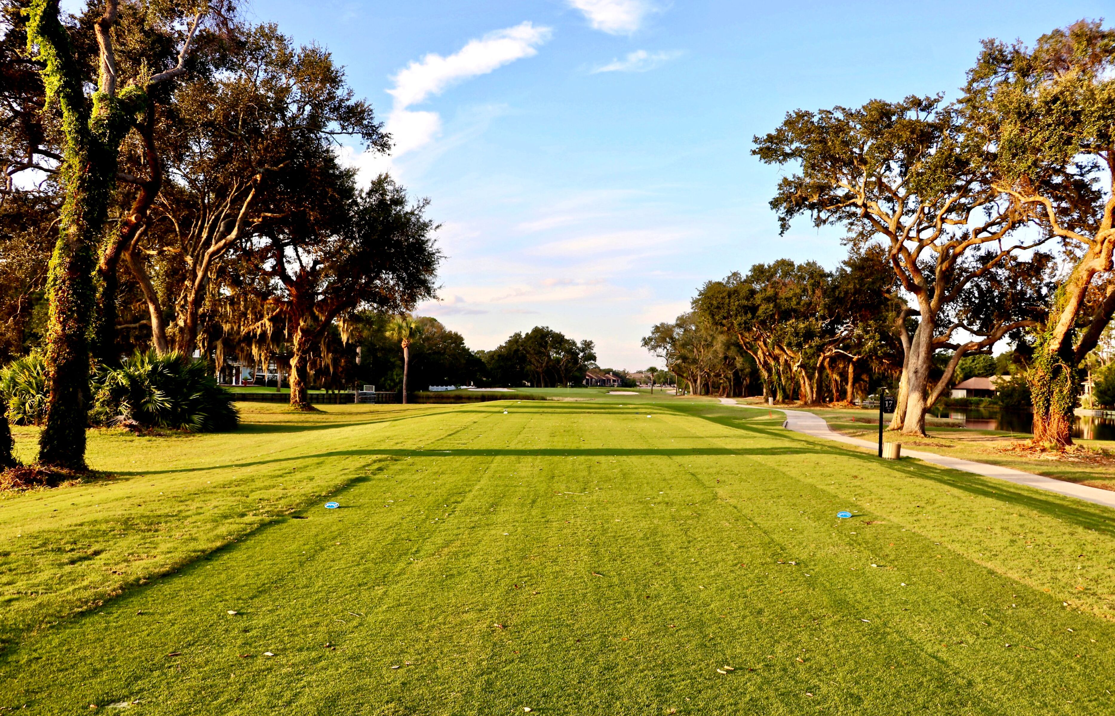A well-maintained golf course with a clear path, surrounded by mature trees and a clear sky.