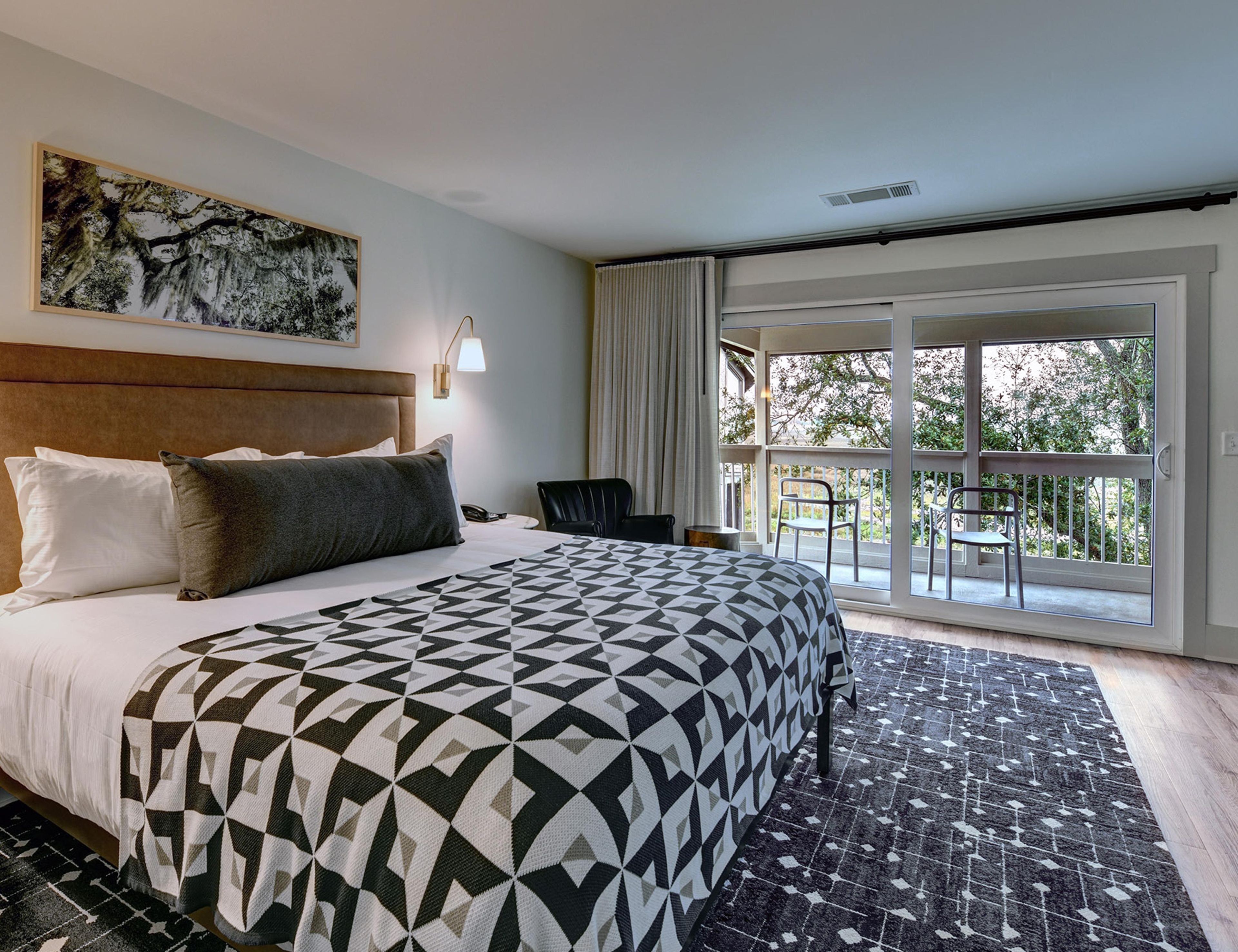 A hotel room with a large bed, a headboard, a nightstand, a desk, a chair, a window with a view of trees, and a patterned rug.