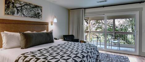 A hotel room with a large bed, a headboard, a nightstand, a desk, a chair, a window with a view of trees, and a patterned rug.