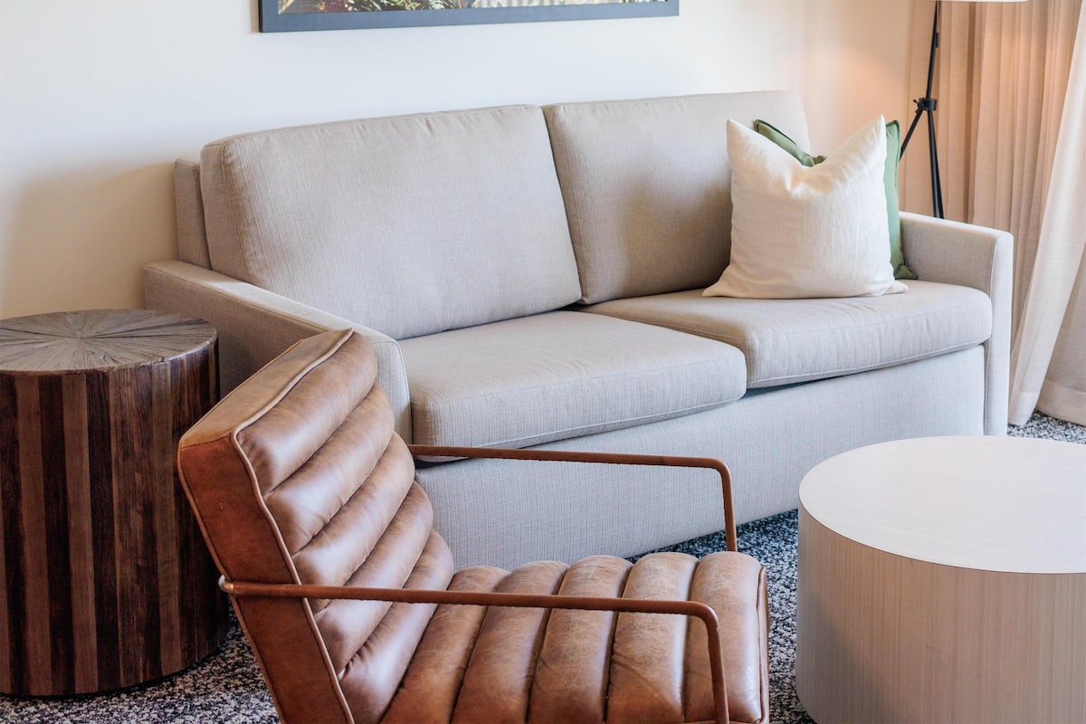 Una habitación de hotel moderna con un sofá gris, un sillón de cuero marrón, una mesita redonda de madera, una lámpara blanca y un cuadro con follaje en la pared.