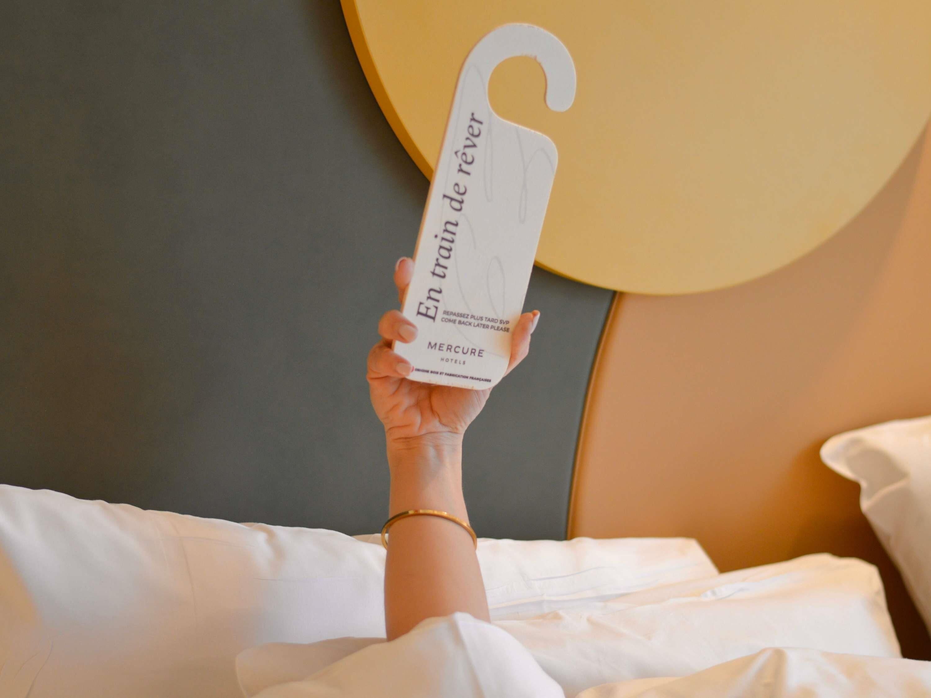 Una mano sostiene una tarjeta blanca con texto en francés, sobre una cama con ropa blanca y un cabecero con diseño circular.