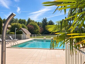 Una piscina con borde de azulejos, rodeada por una valla blanca, y un edificio con techo inclinado al fondo.