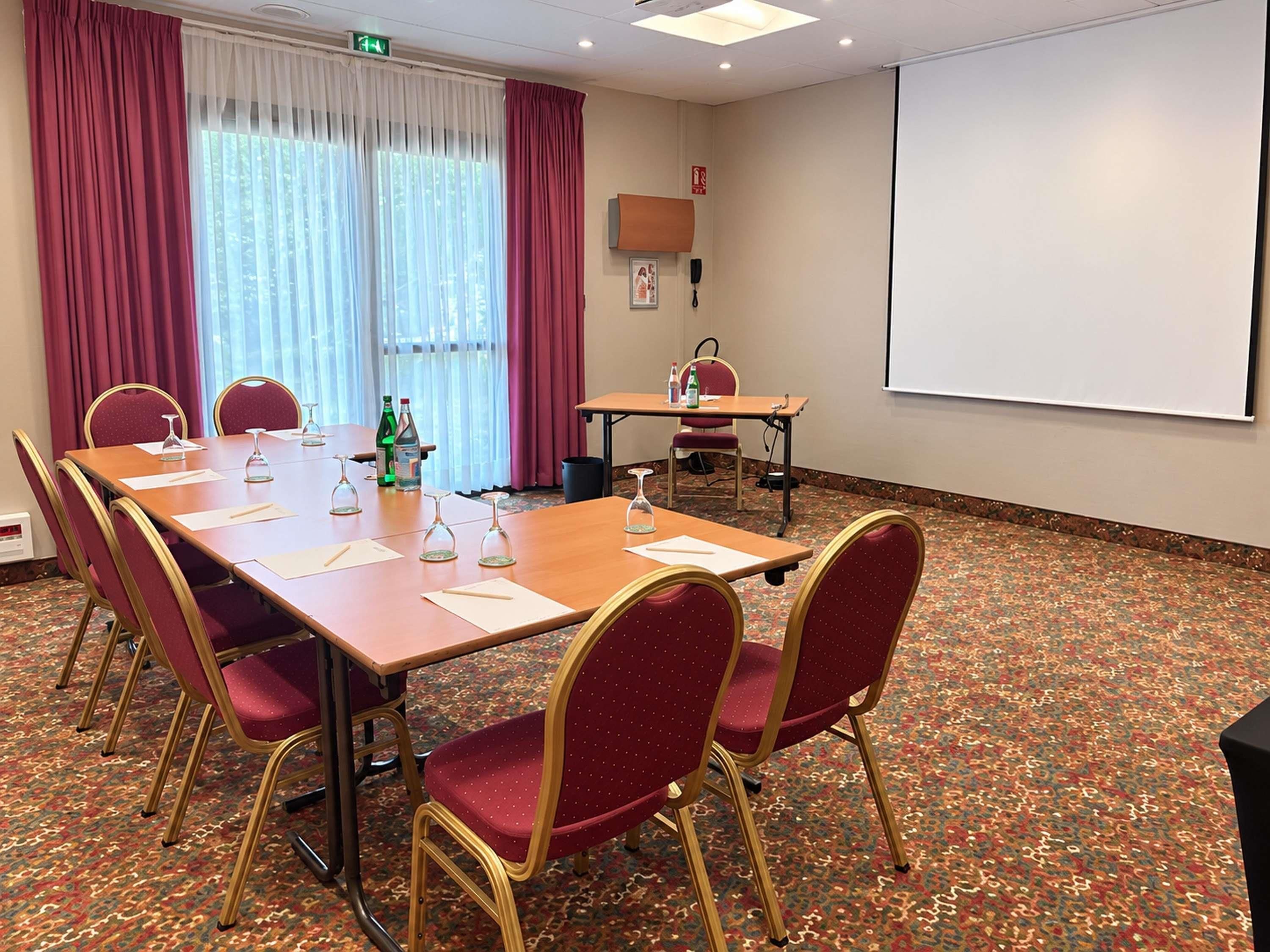 Una sala de conferències amb una taula llarga, cadires vermelles, una pantalla de projector i un sòl amb moqueta.
