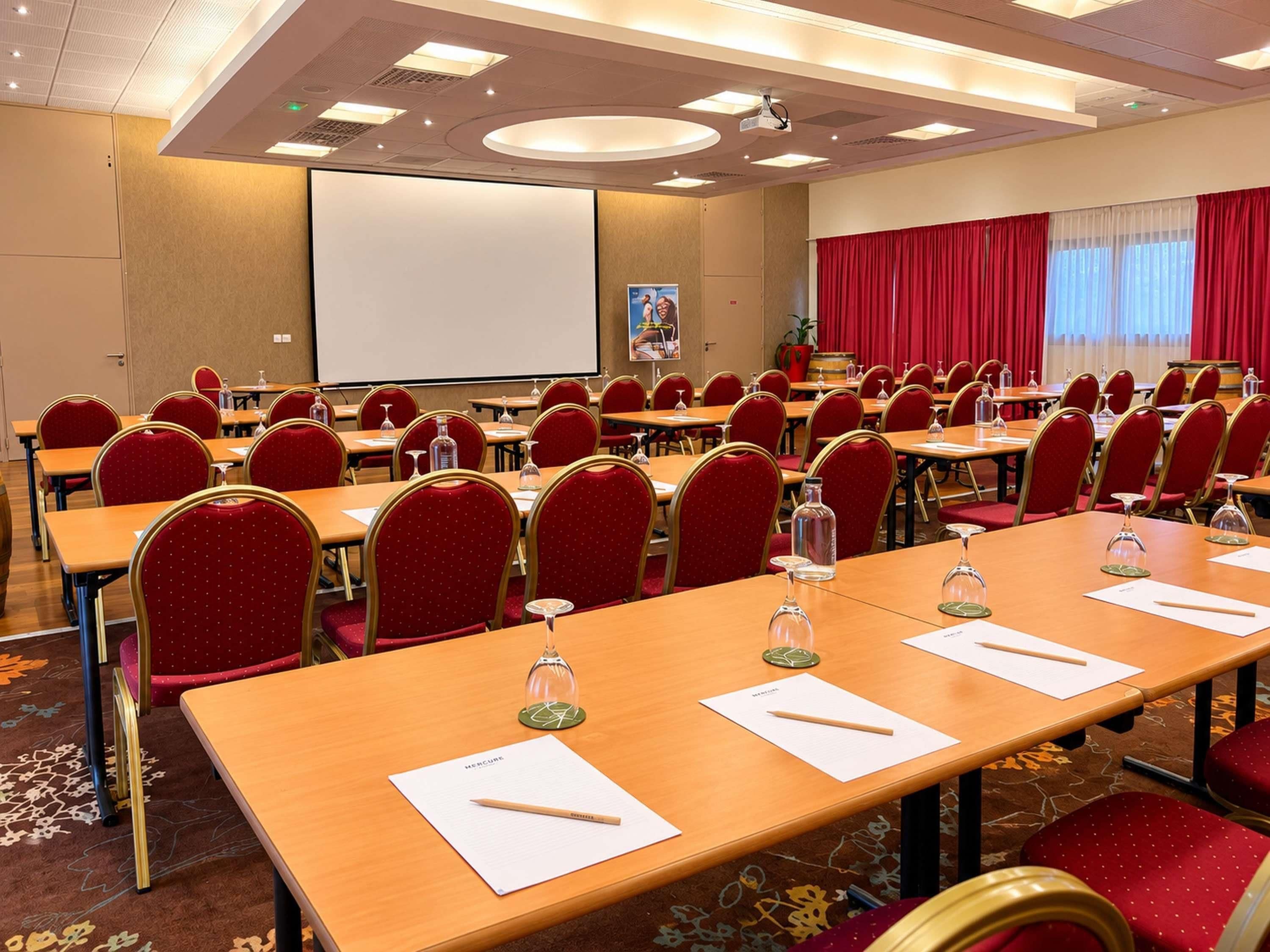 Ein Konferenzraum mit Tischen, Stühlen, einer Projektionsleinwand und einem großen Fenster mit Vorhängen.