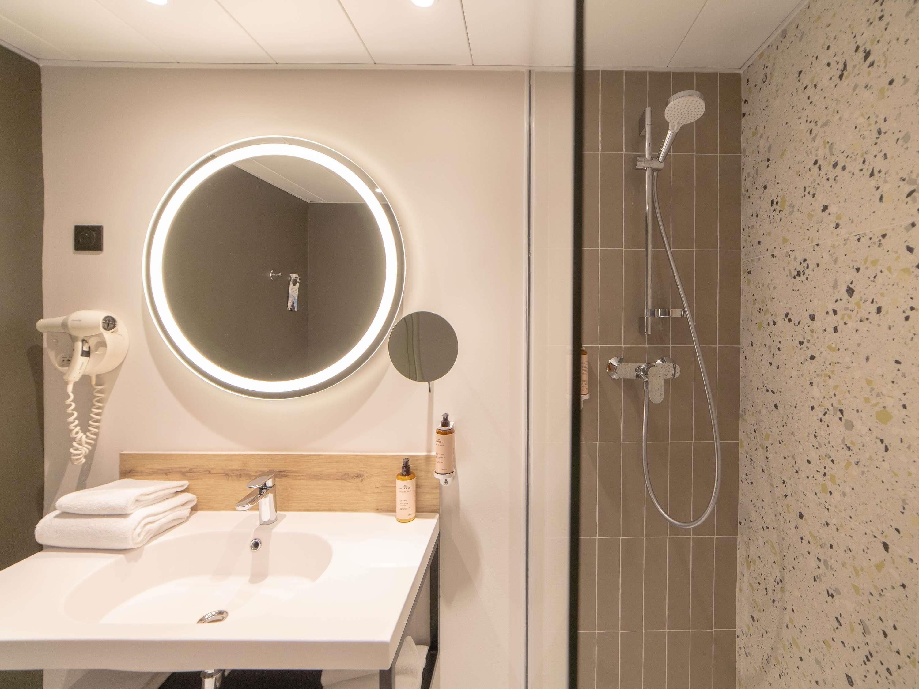 Un bagno con uno specchio rotondo, un asciugacapelli, un lavandino e una doccia.