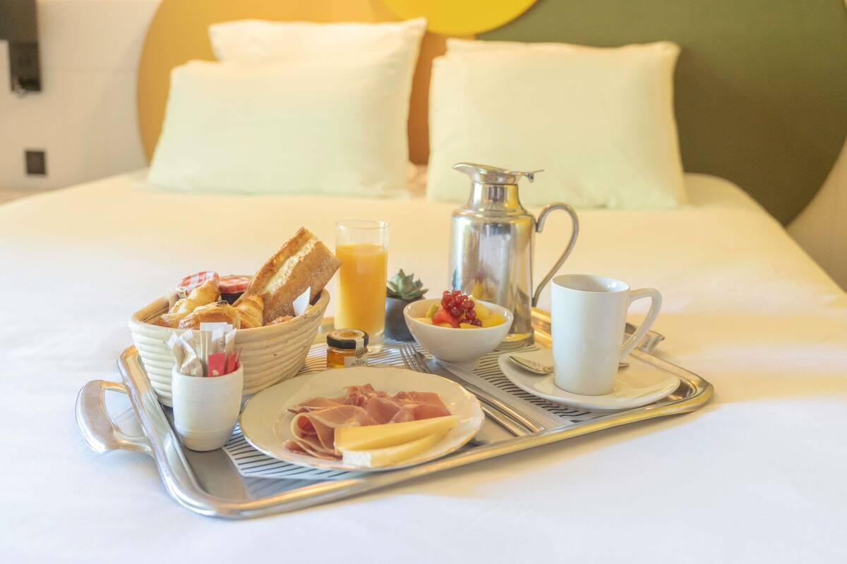 Ein Frühstückswagen mit Brot, Obst, Käse und Schinken auf einem Bett mit weißer Bettwäsche und einem grünen Kopfteil.