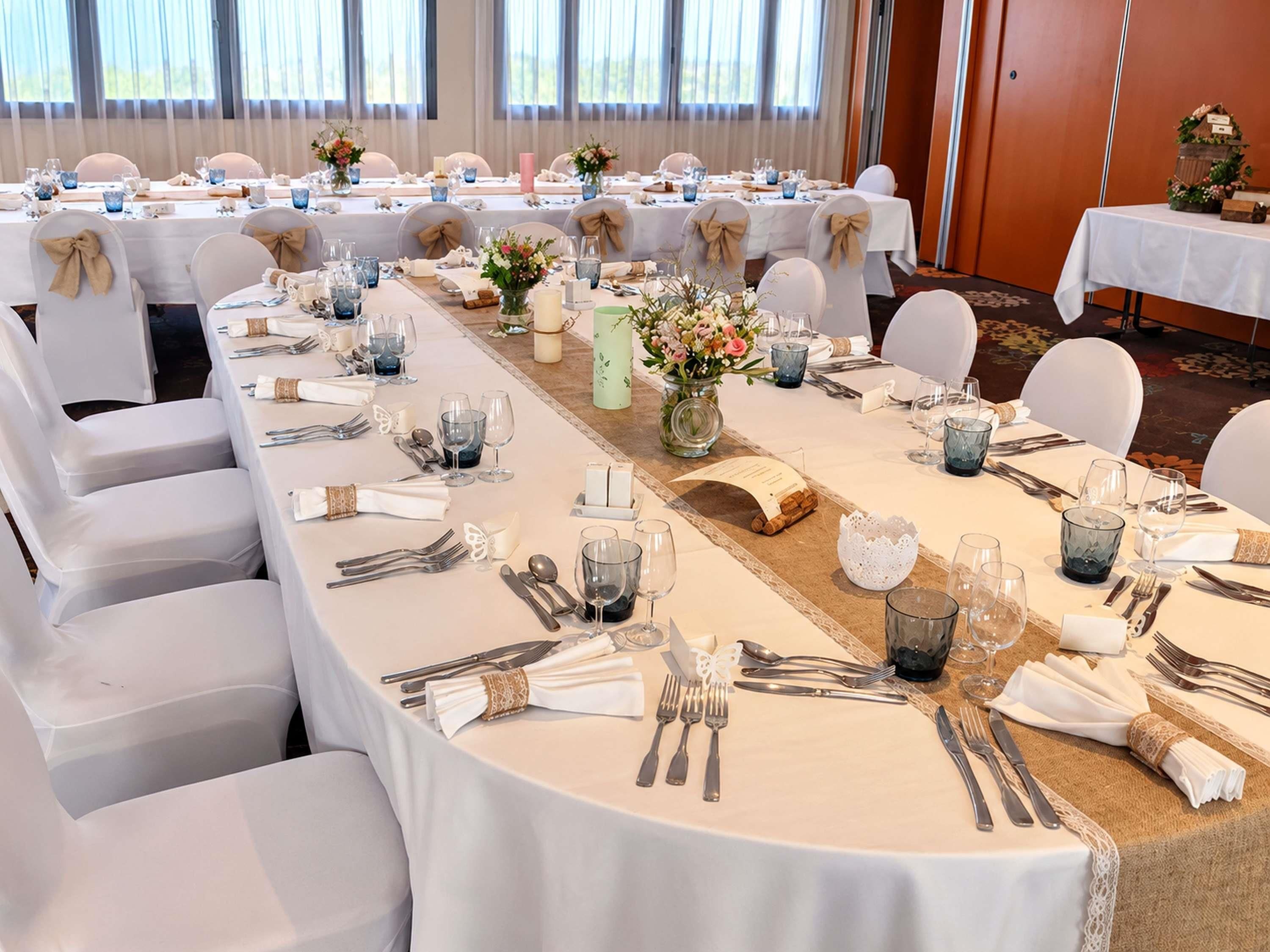 Une salle de banquet avec des tables rondes dressées pour un événement formel, agrémentée de nappes blanches, de couverts disposés avec soin et de centres de table floraux.