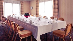 Un salón de conferencias con una larga mesa dispuesta para una reunión, rodeado de sillas, con ventanas y cortinas.