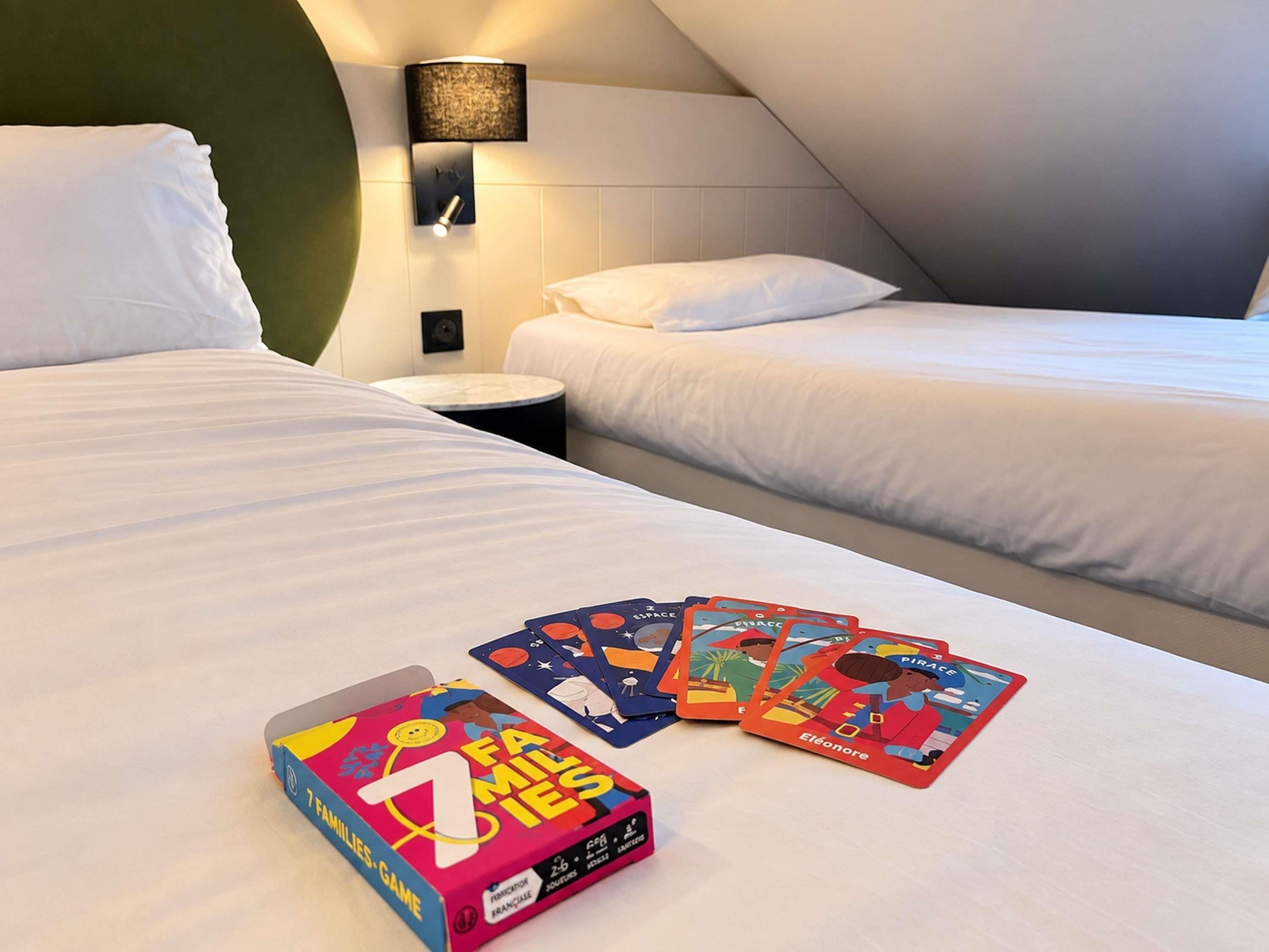 A hotel room with two beds, a nightstand, and a lamp. There is a game box labeled '7 Families' and some game cards on the bed.
