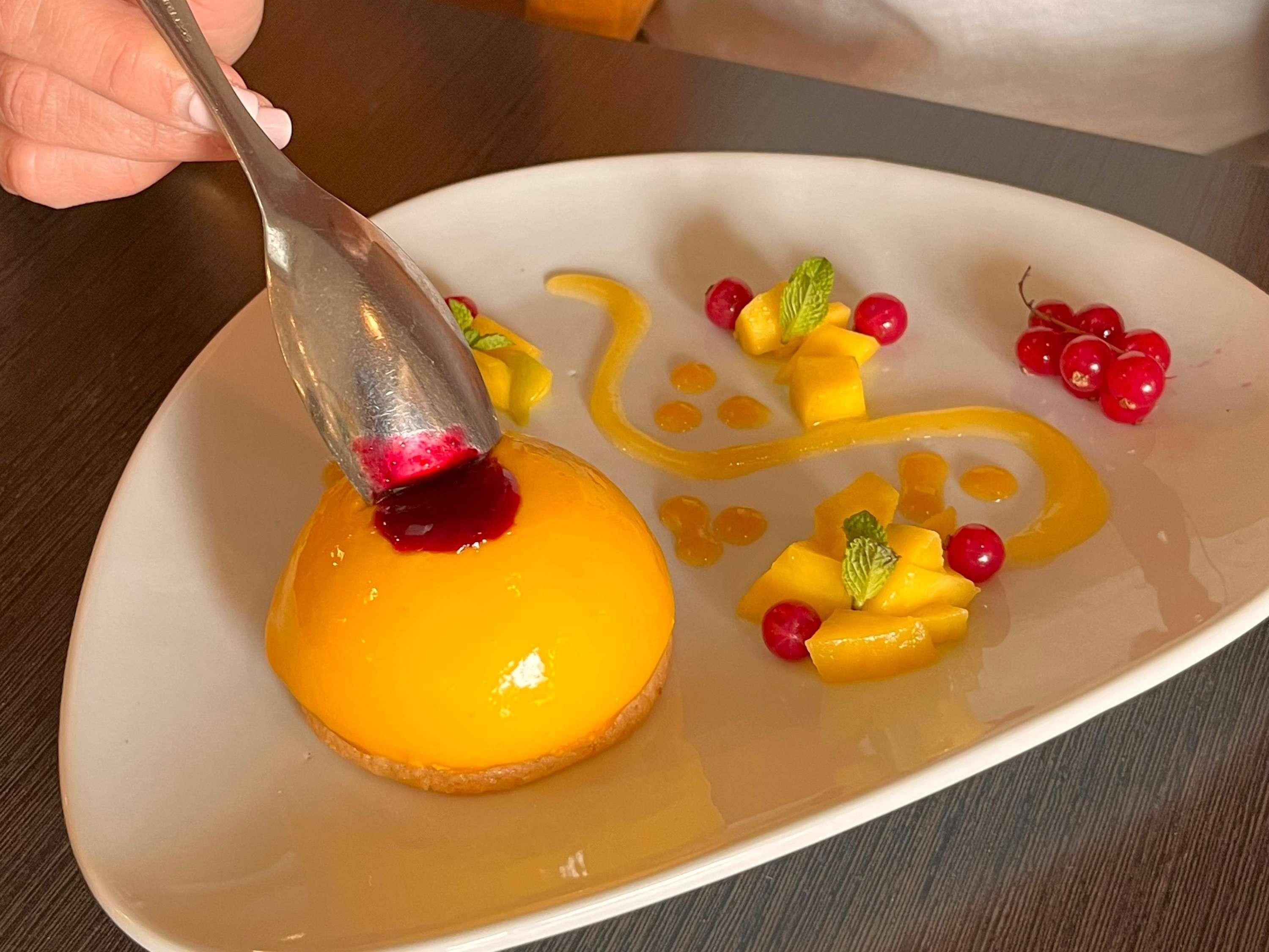 Une assiette de dessert surmontée d’une garniture lisse et brillante, décorée de dés de mangue, de baies rouges et de feuilles de menthe.