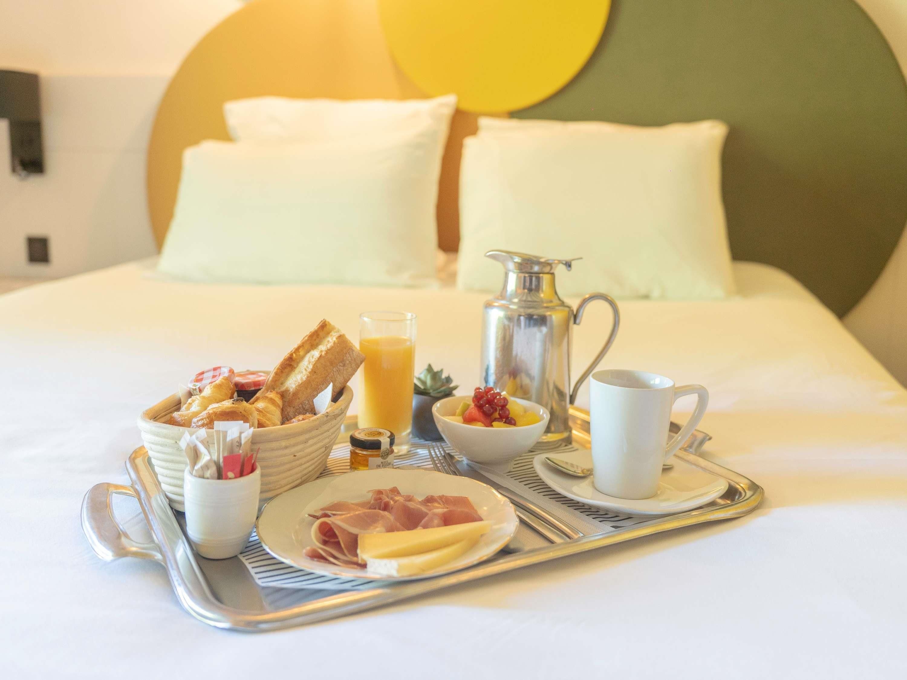 Ein Frühstücksbrett mit Brot, Obst, Käse und Schinken auf einem Bett mit Kissen und einer Metallkanne.