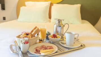 Ein Frühstücksbrett mit Brot, Obst, Käse und Schinken auf einem Bett mit Kissen und einer Metallkanne.