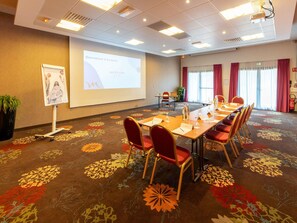 Une salle de conférence avec une longue table, des chaises rouges, un écran de projection et une moquette à motifs floraux.