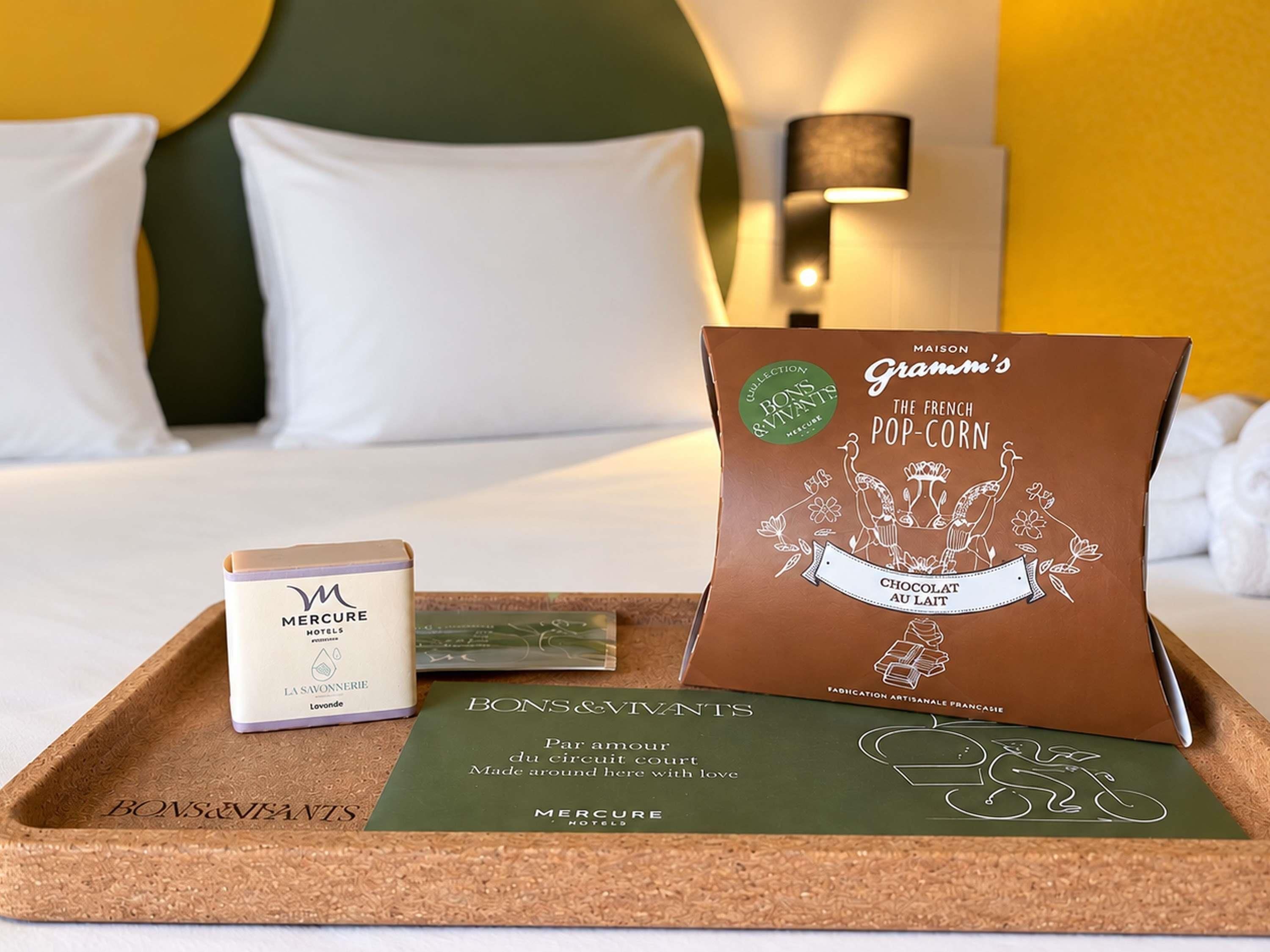 A hotel room with a bed, pillows, a tray with a chocolate bar, a box of Mercure hotel toiletries, and a Bons & Vivants brochure.