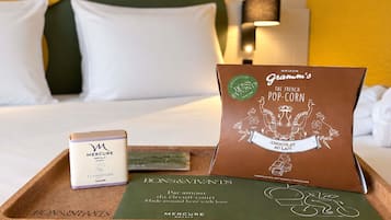 A hotel room with a bed, pillows, a tray with a chocolate bar, a box of Mercure hotel toiletries, and a Bons & Vivants brochure.