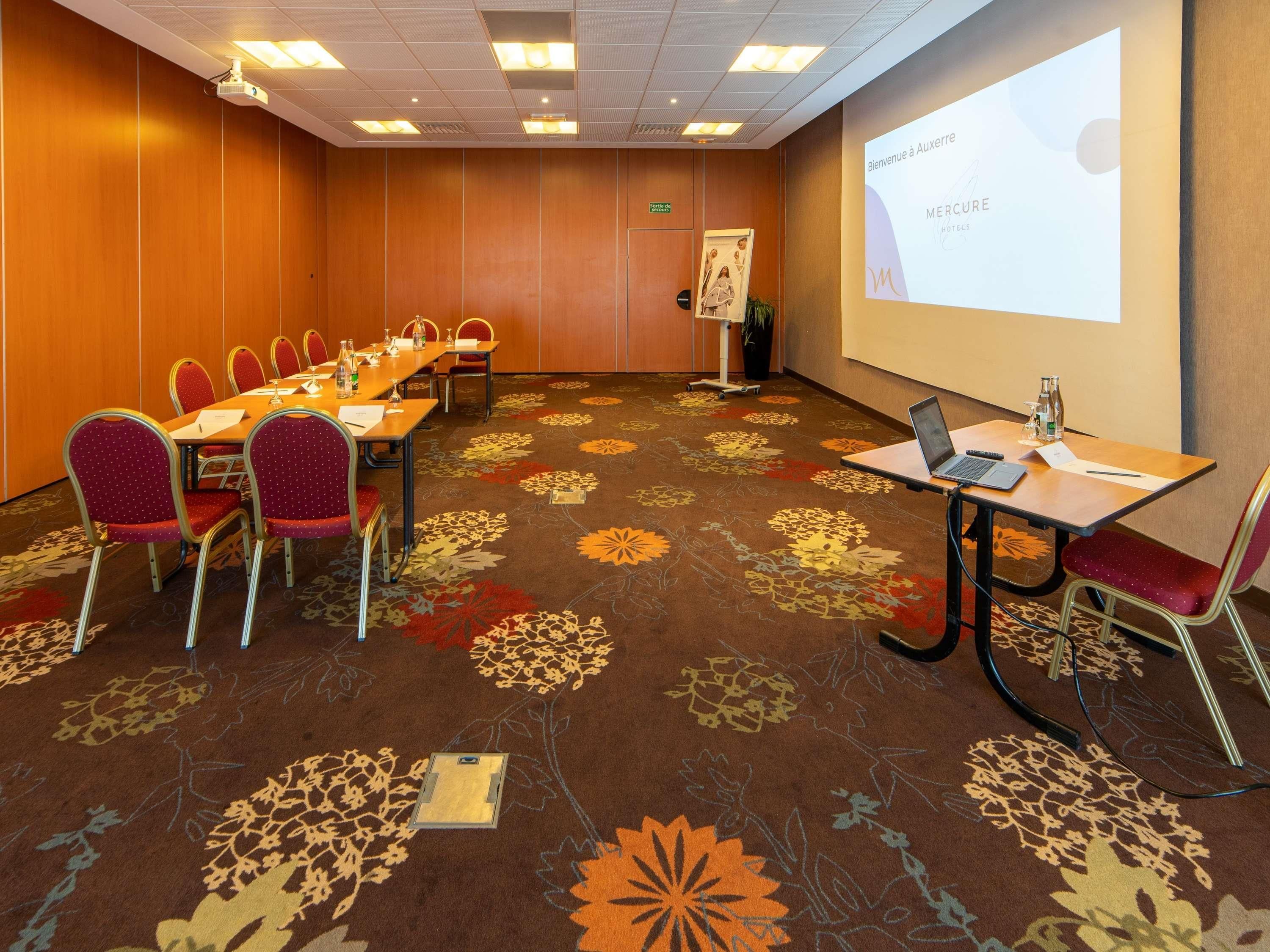 A conference room with a long table, red chairs, a projector screen, and a floral carpet.