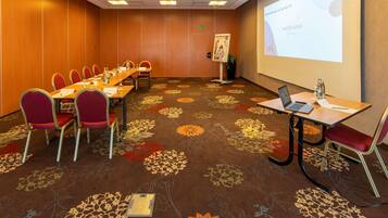 A conference room with a long table, red chairs, a projector screen, and a floral carpet.