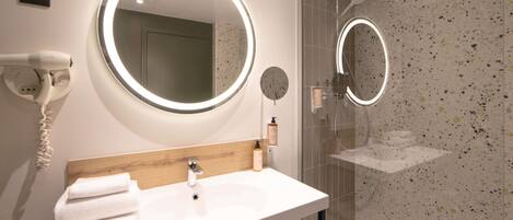 A modern bathroom with a white sink, a round mirror, a handheld hair dryer, and a glass-enclosed shower.