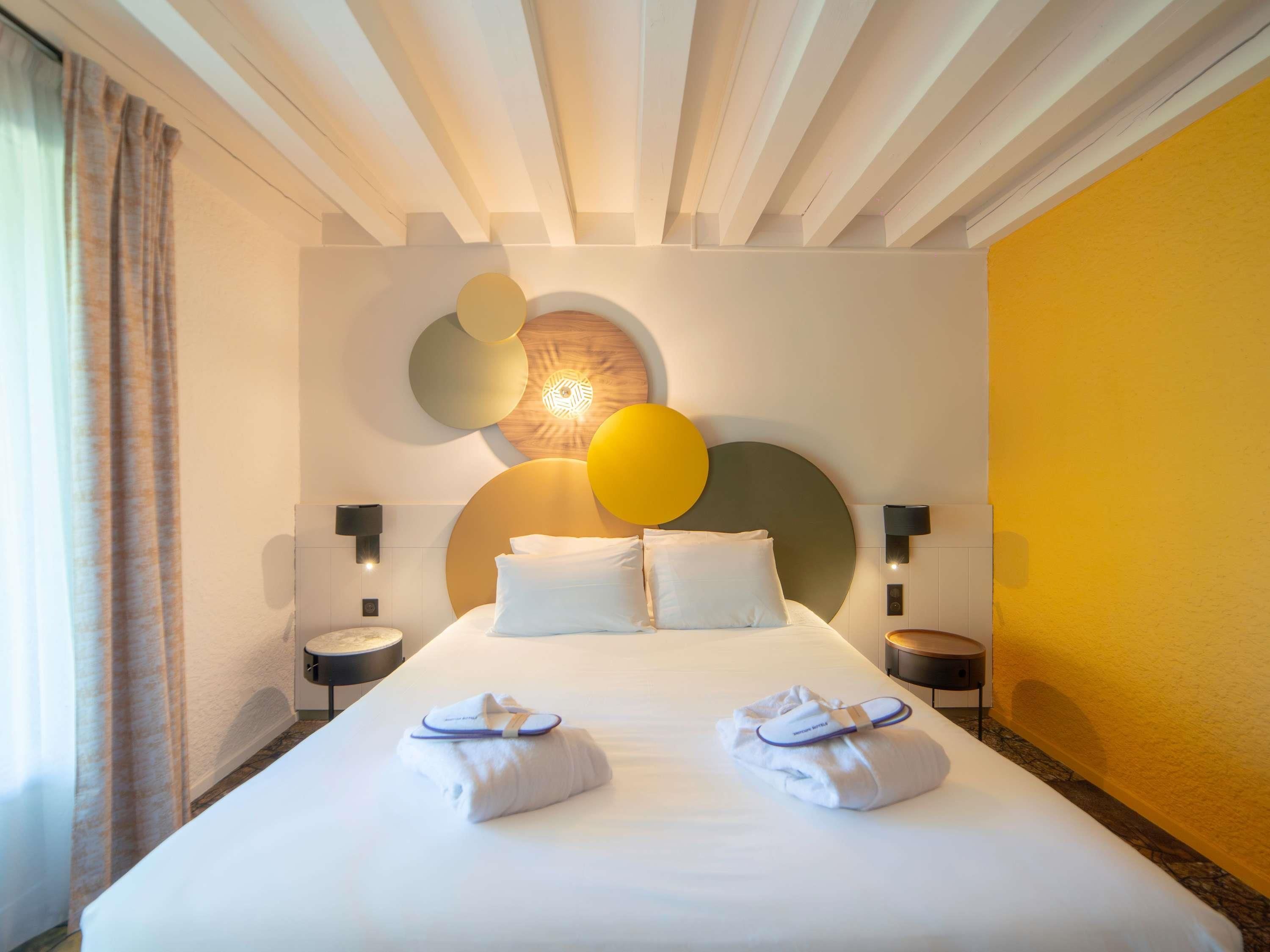 Habitación de hotel con una cama grande, dos mesitas de noche, una lámpara de pared y una pared de acento amarilla.