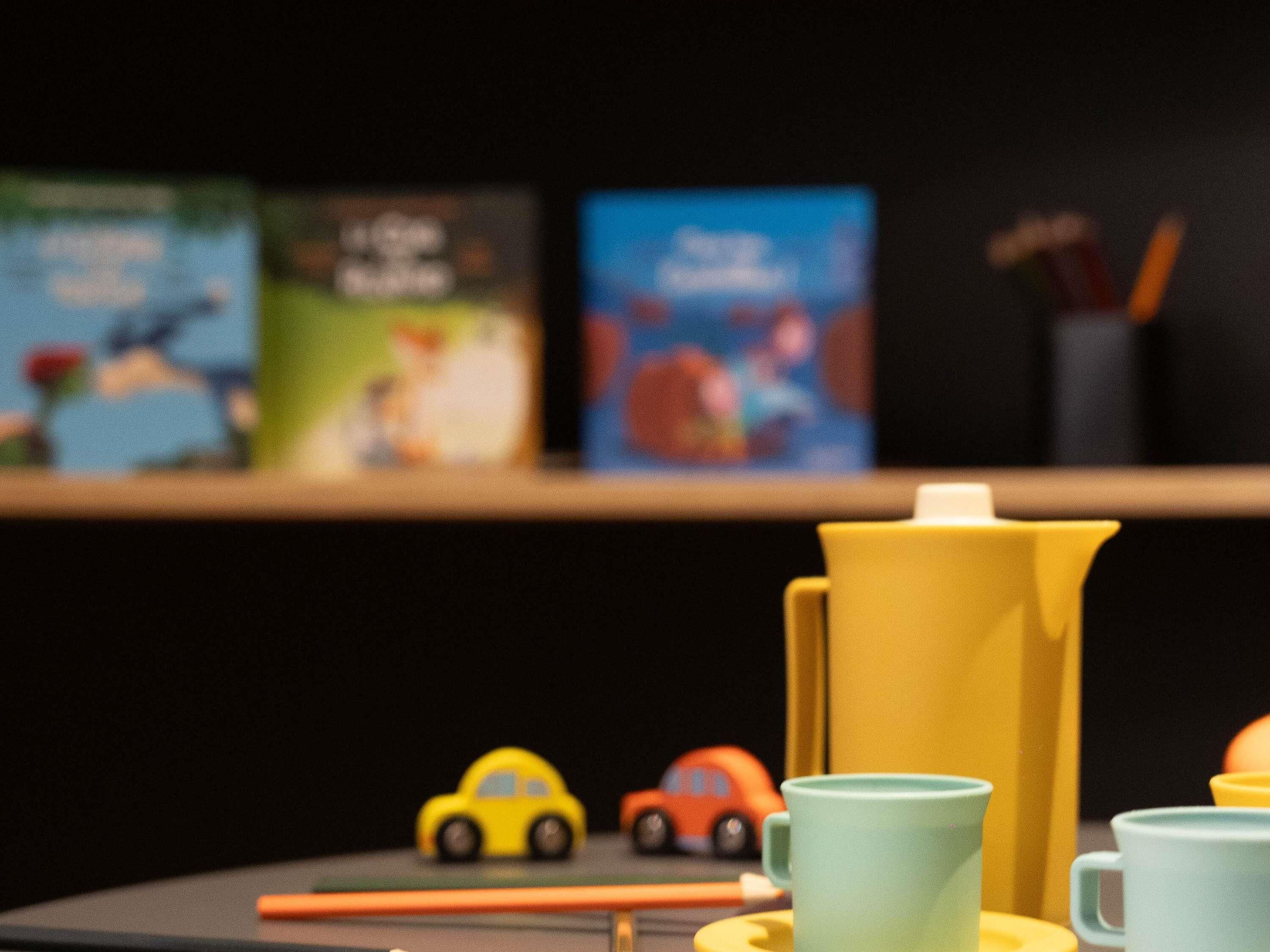 Een plank met boeken en een geel object, mogelijk een bak, mét speelgoedauto's ervoor.