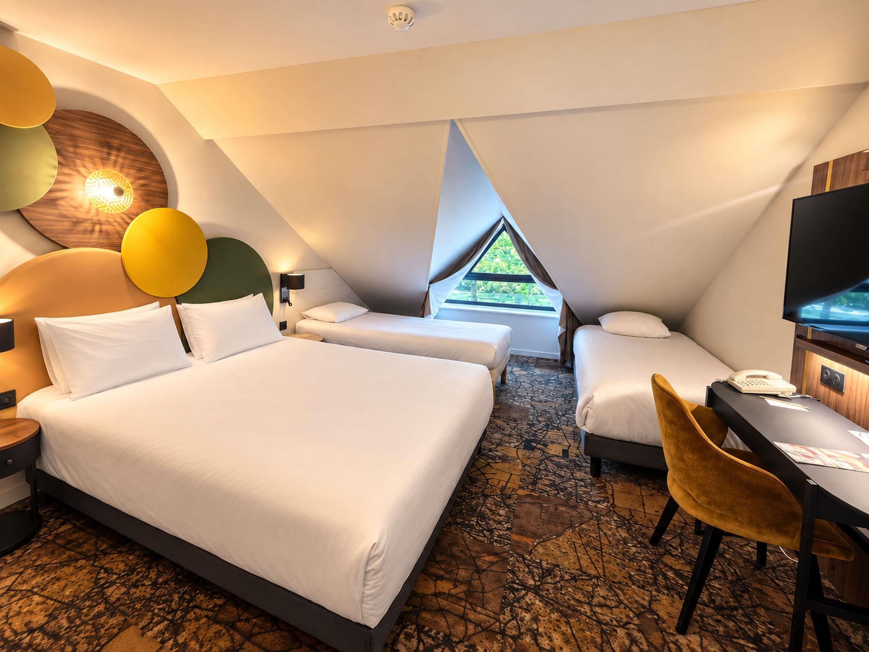 A modern hotel room with a large bed, two smaller beds, a desk with a chair, and a television.