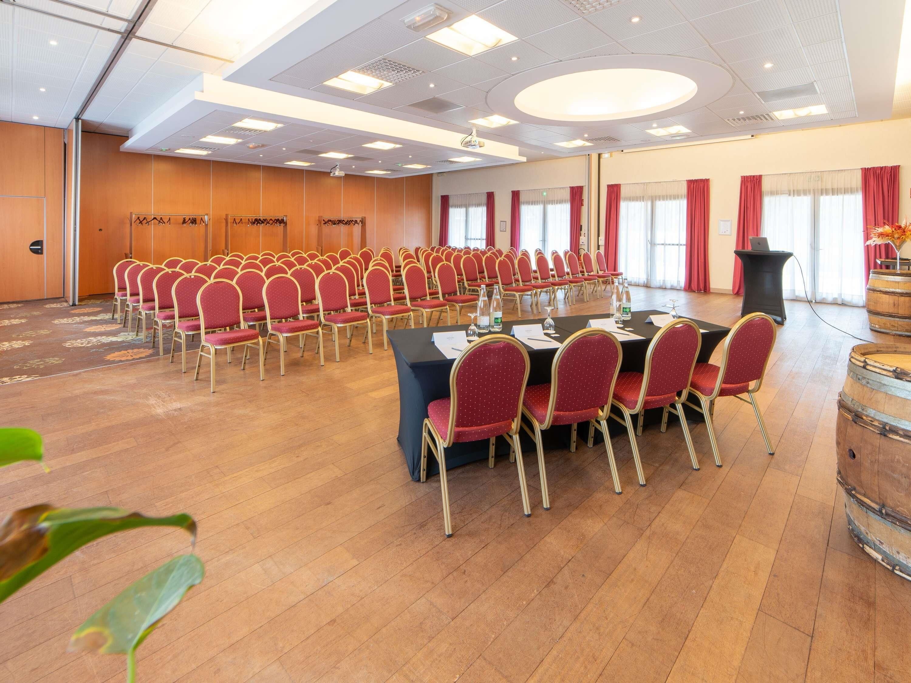 A conference room with rows of red chairs, a table with papers, and a wooden floor.