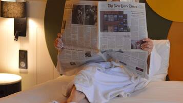 A person reading The New York Times on a bed with a lamp and a wall socket visible.