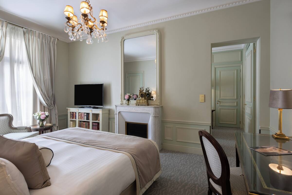 A hotel room with a bed, a television, a fireplace, a mirror, a desk with a lamp, and a chair.