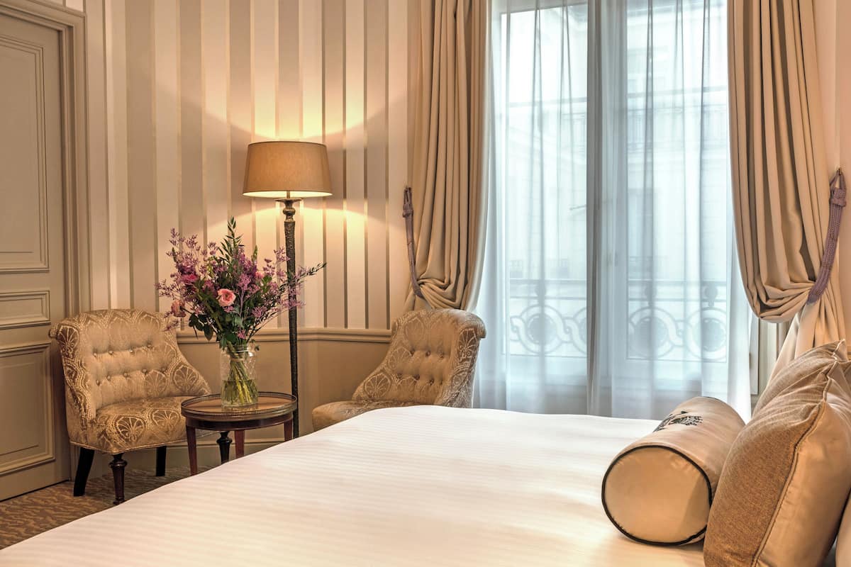 A hotel room with a large bed, two armchairs, a small table with a lamp, and a vase of flowers.