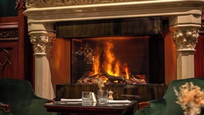 Ein Kamin mit Feuer, ein Tisch mit Gläsern und einer Vase sowie grüne Polsterstühle.