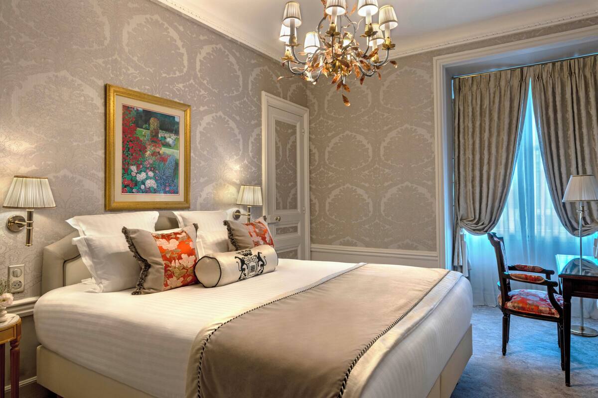 A hotel room with a large bed, two bedside lamps, a chandelier, a framed painting, and a view of the outdoors through a window with curtains.