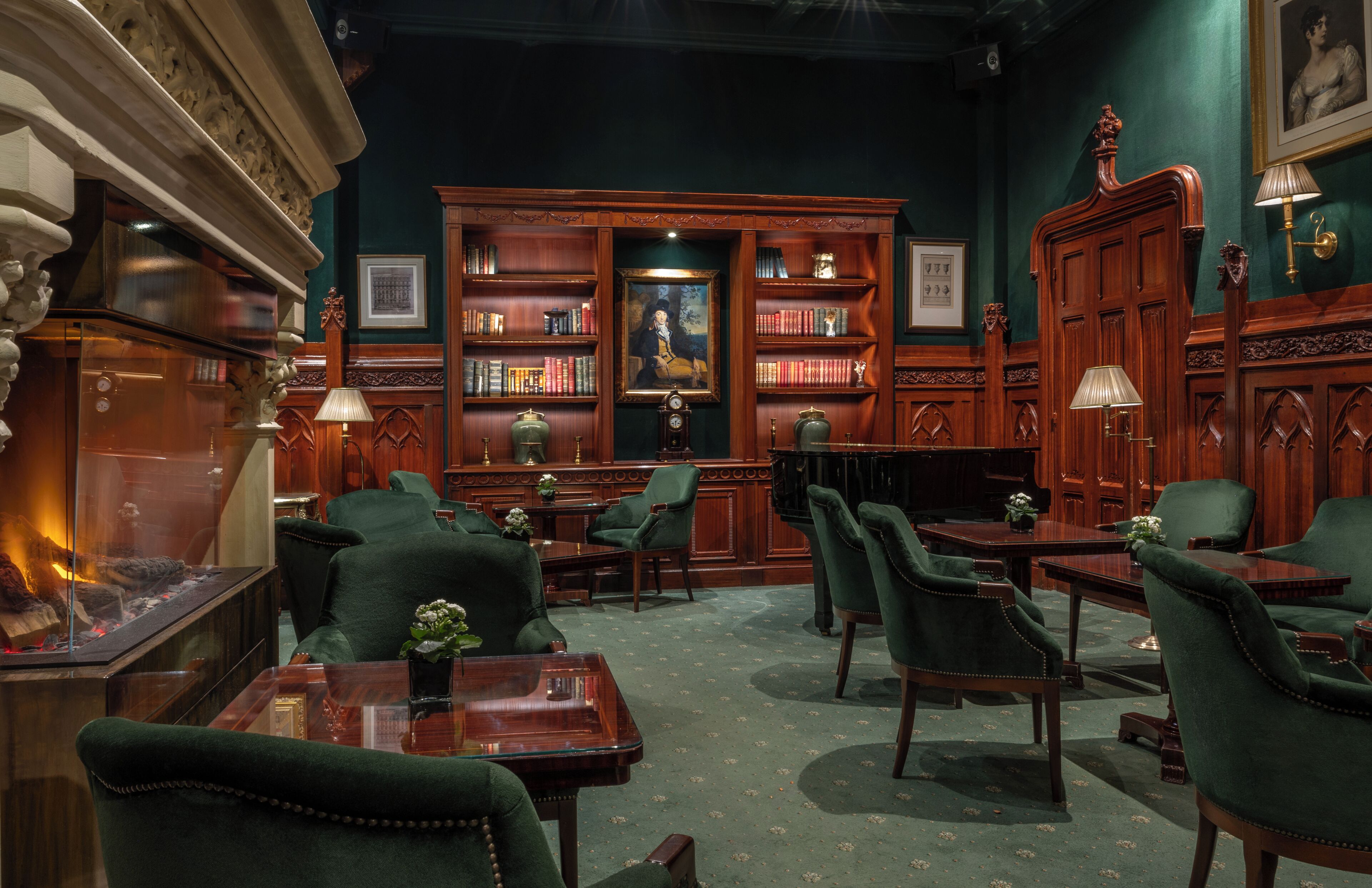 A library with bookshelves, green upholstered chairs, a fireplace, and a desk with a lamp.