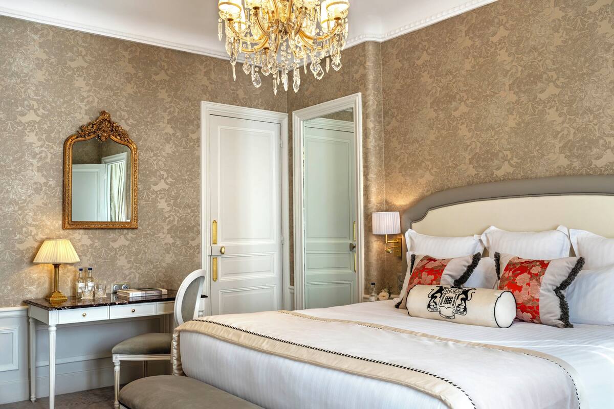 A bedroom with a bed, a desk, a chair, a mirror, and a chandelier.