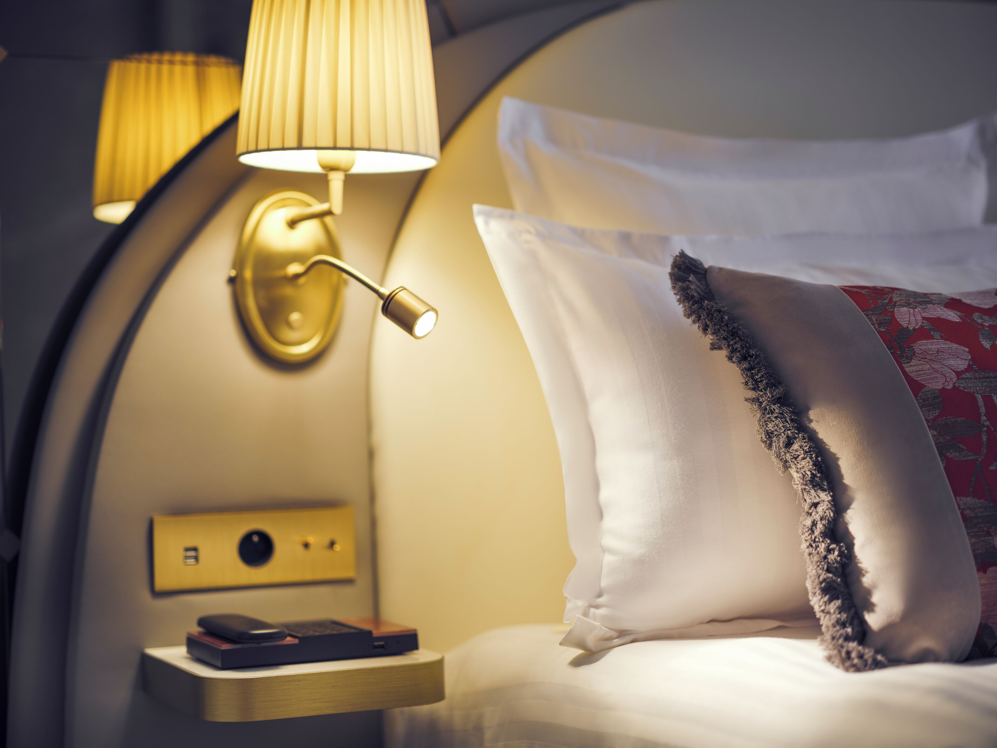 Ein ordentlich bezogenes Bett mit weißen Leinen und einem Zierkissen, ein Nachttisch mit Lampe und eine wandmontierte Bedieneinheit.