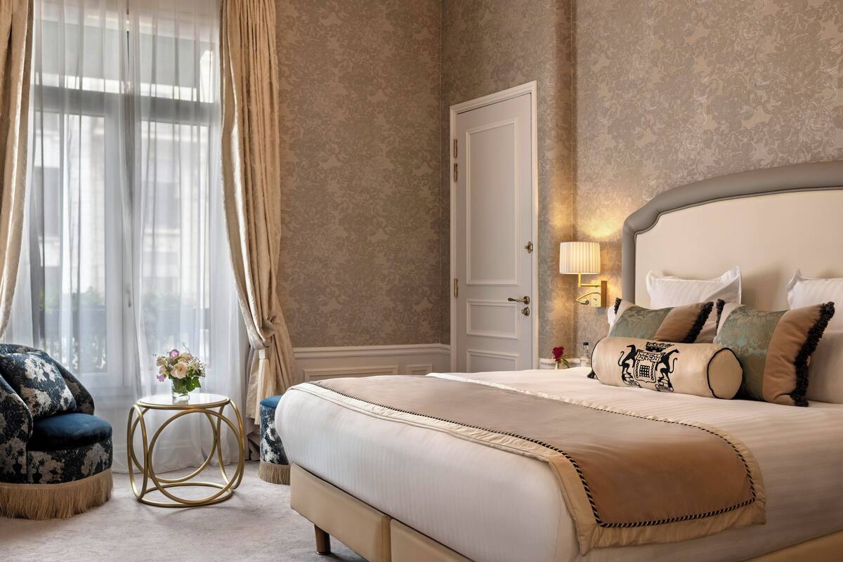 A bedroom with a large bed, a chair, a small round table with a vase of flowers, and a door.