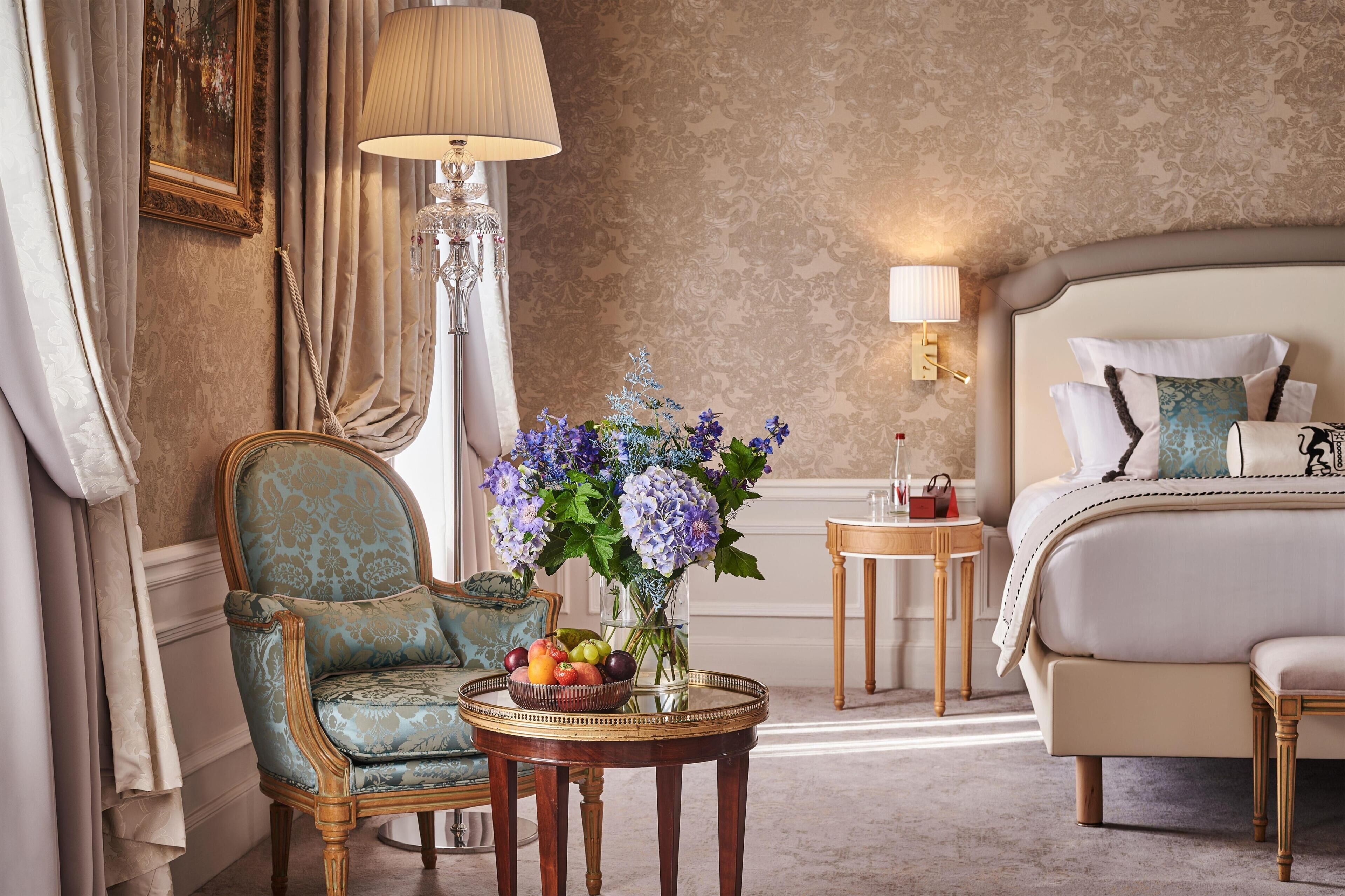 Habitación de hotel con cama, una silla, una mesa con fruta y flores, una lámpara y un cuadro en la pared.