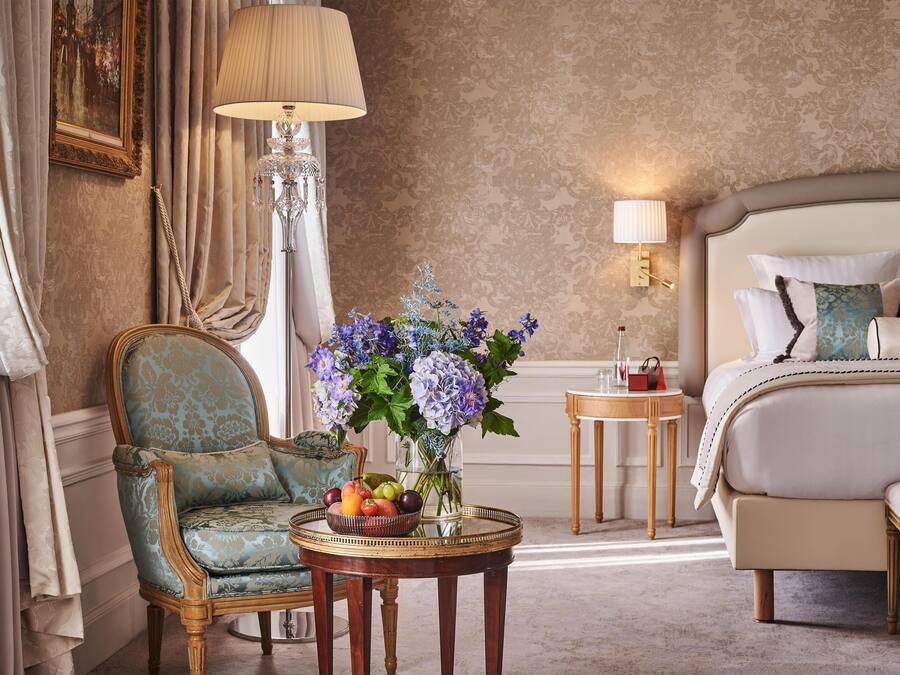 Habitación de hotel con cama, una silla, una mesa con fruta y flores, una lámpara y un cuadro en la pared.