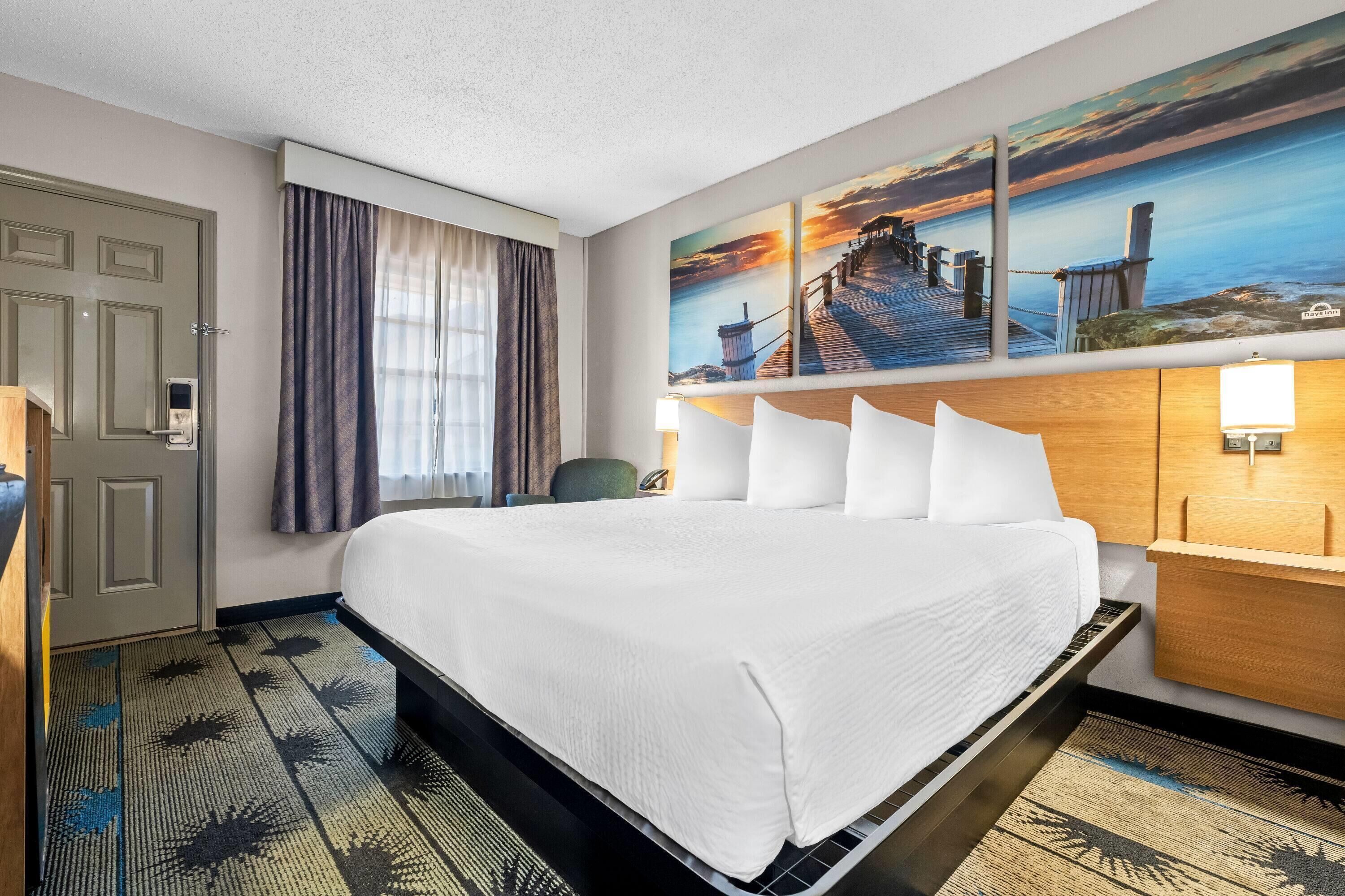 Habitación de hotel con una cama grande, cabecera de madera, mesita de noche con lámpara, un cuadro de un muelle y una puerta con manija.